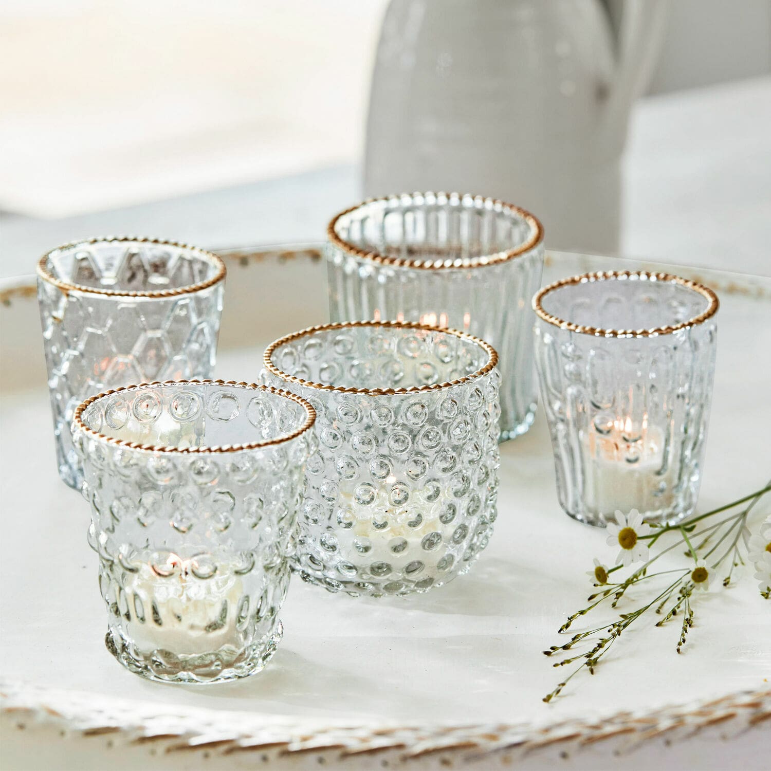 Set de 3 farolillos Jarnac Conjunto de cinco portavelas de vidrio con borde dorado y textura, sobre bandeja blanca decorativa