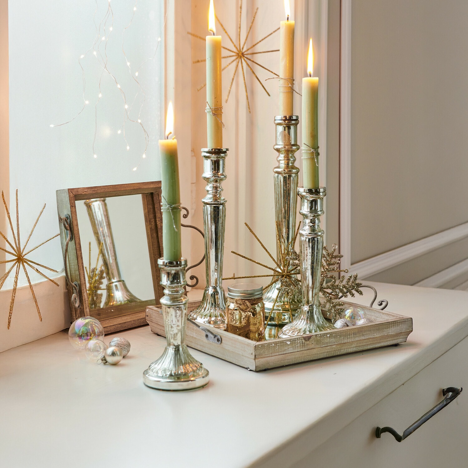 Set de 4 candeleros Selby Candelabros de metal plateado con velas verdes y crema sobre bandeja decorativa de madera
