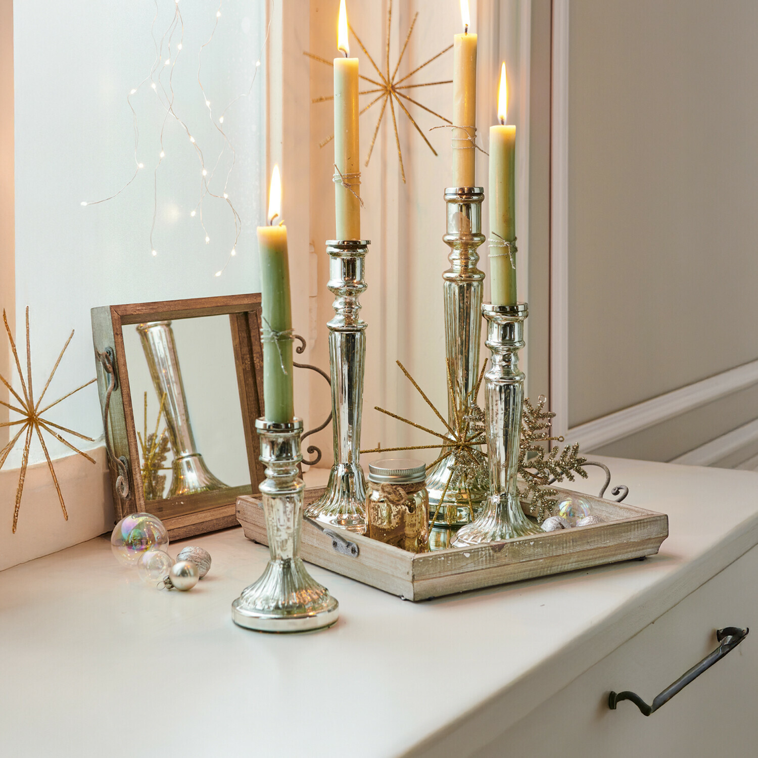 Set de 4 candeleros Selby Candelabros de metal plateado con velas verdes y crema sobre bandeja decorativa de madera