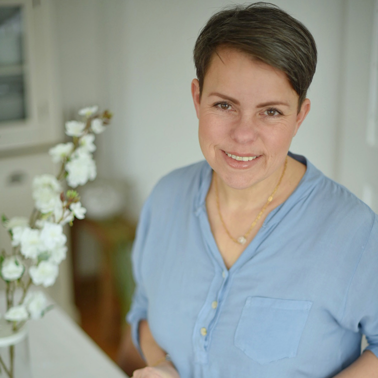 Mujer sonriente con blusa azul junto a flores blancas en un interior luminoso