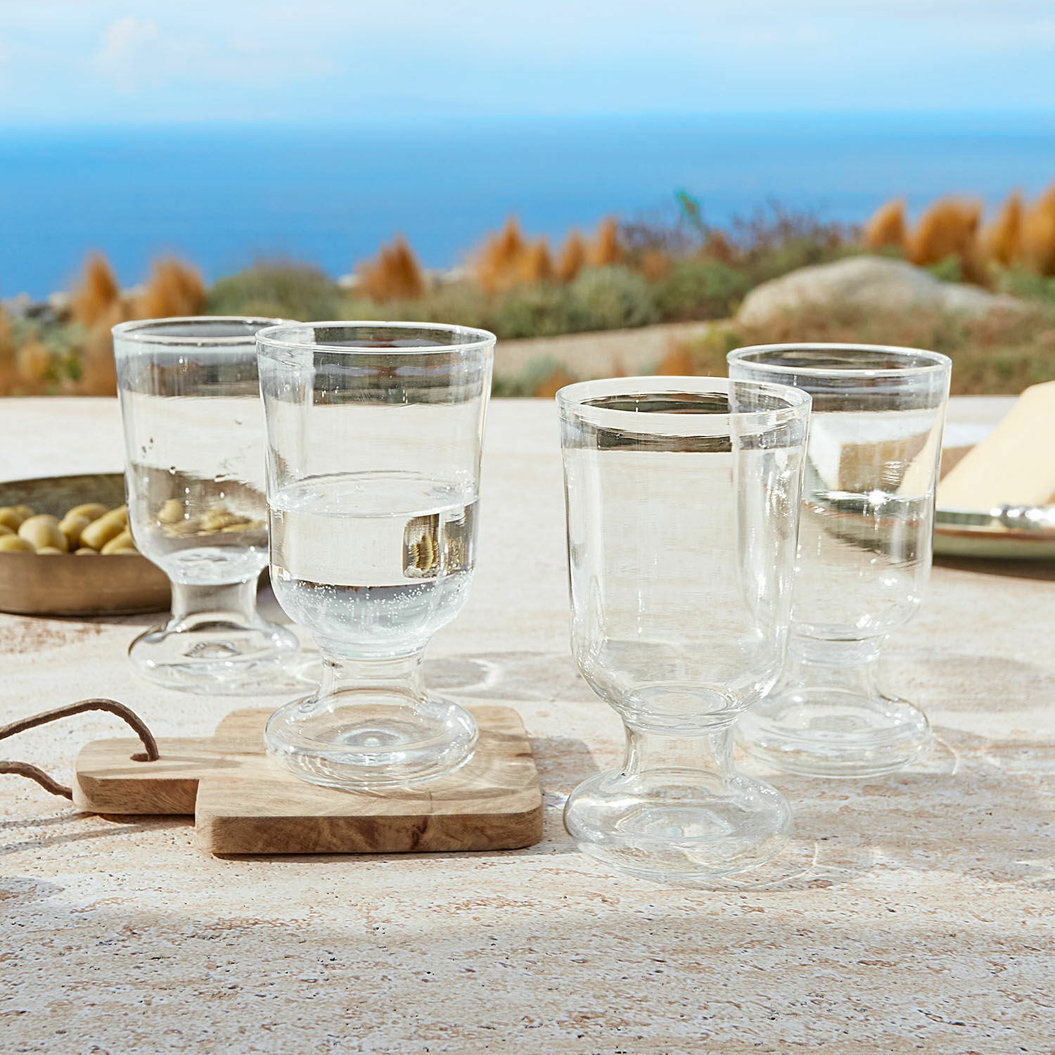Set de 4 vasos Yvoncourt Conjunto de cuatro vasos transparentes con pie sobre mesa con vista al mar