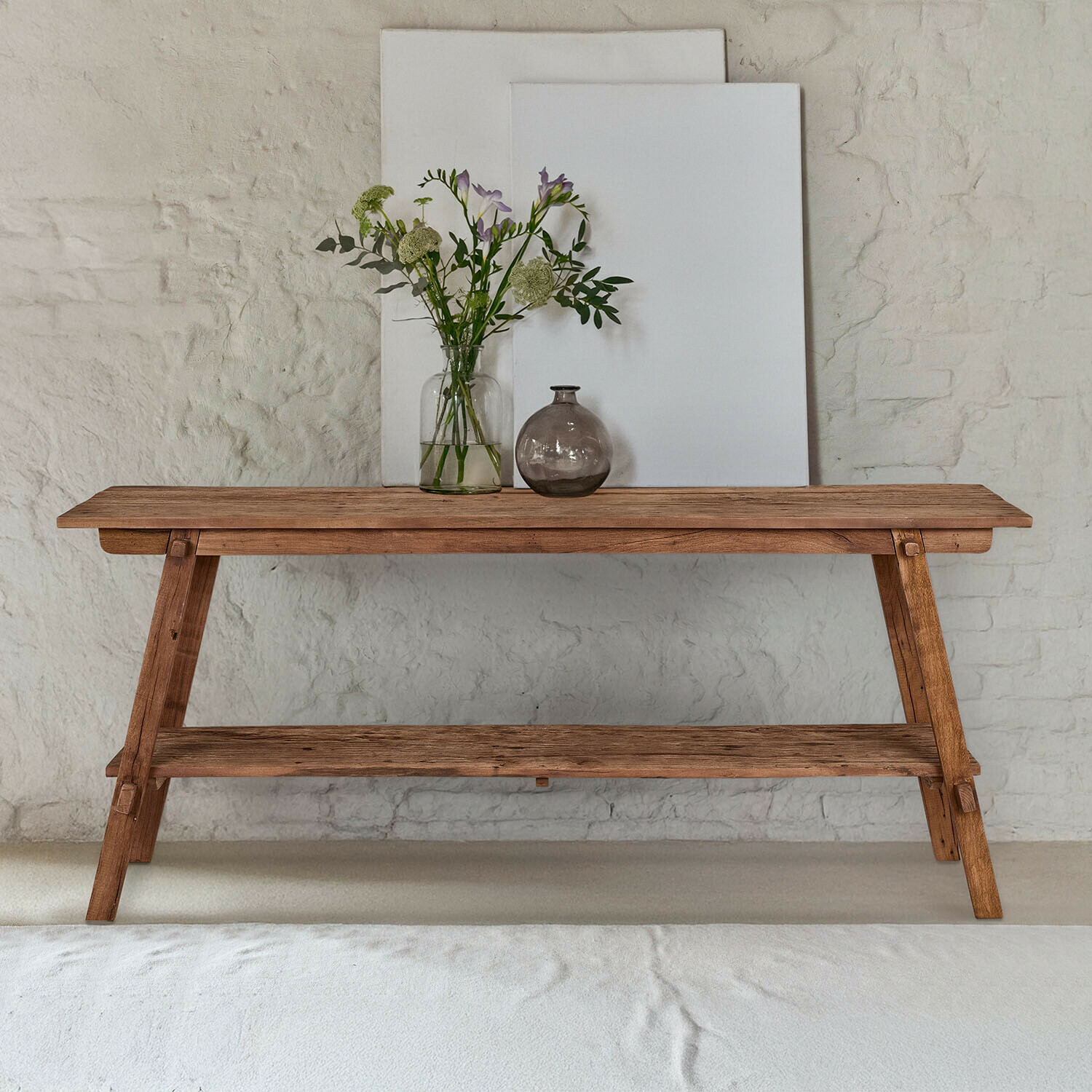 Mesa de madera natural con estante inferior, decorada con jarrones y flores frente a pared blanca