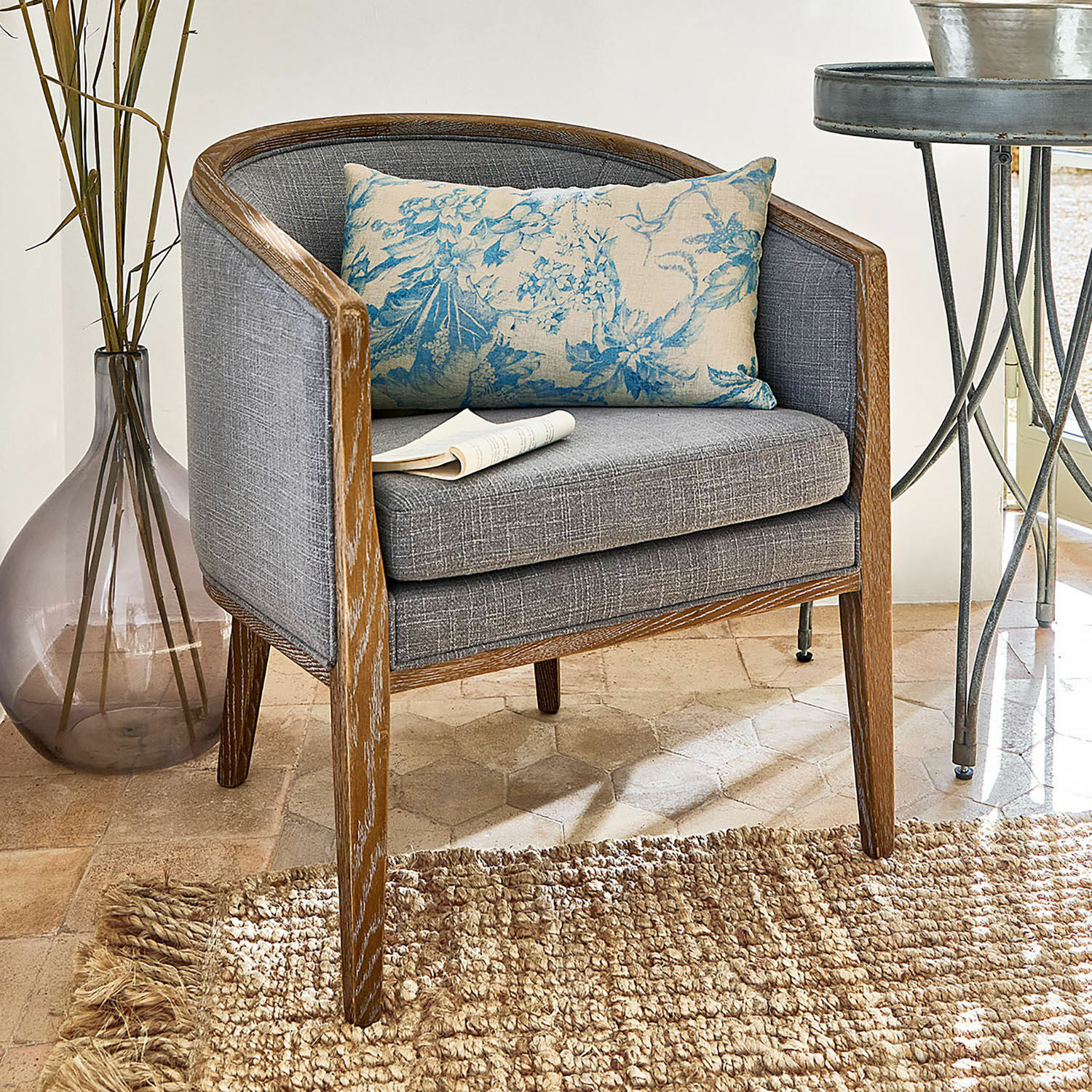 Sillón gris con marco de madera y cojín azul floral sobre alfombra natural tejida