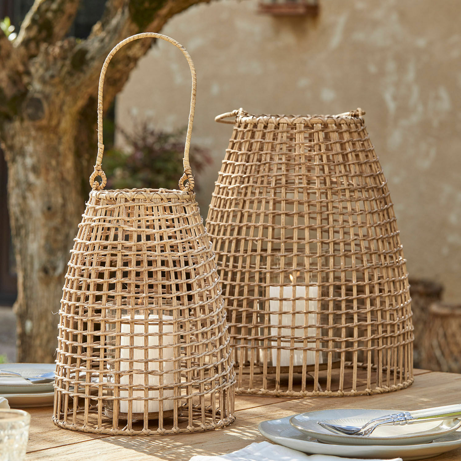 Set de 2 linternas Hillendale Dos faroles de ratán natural con velas blancas sobre una mesa de madera al aire libre