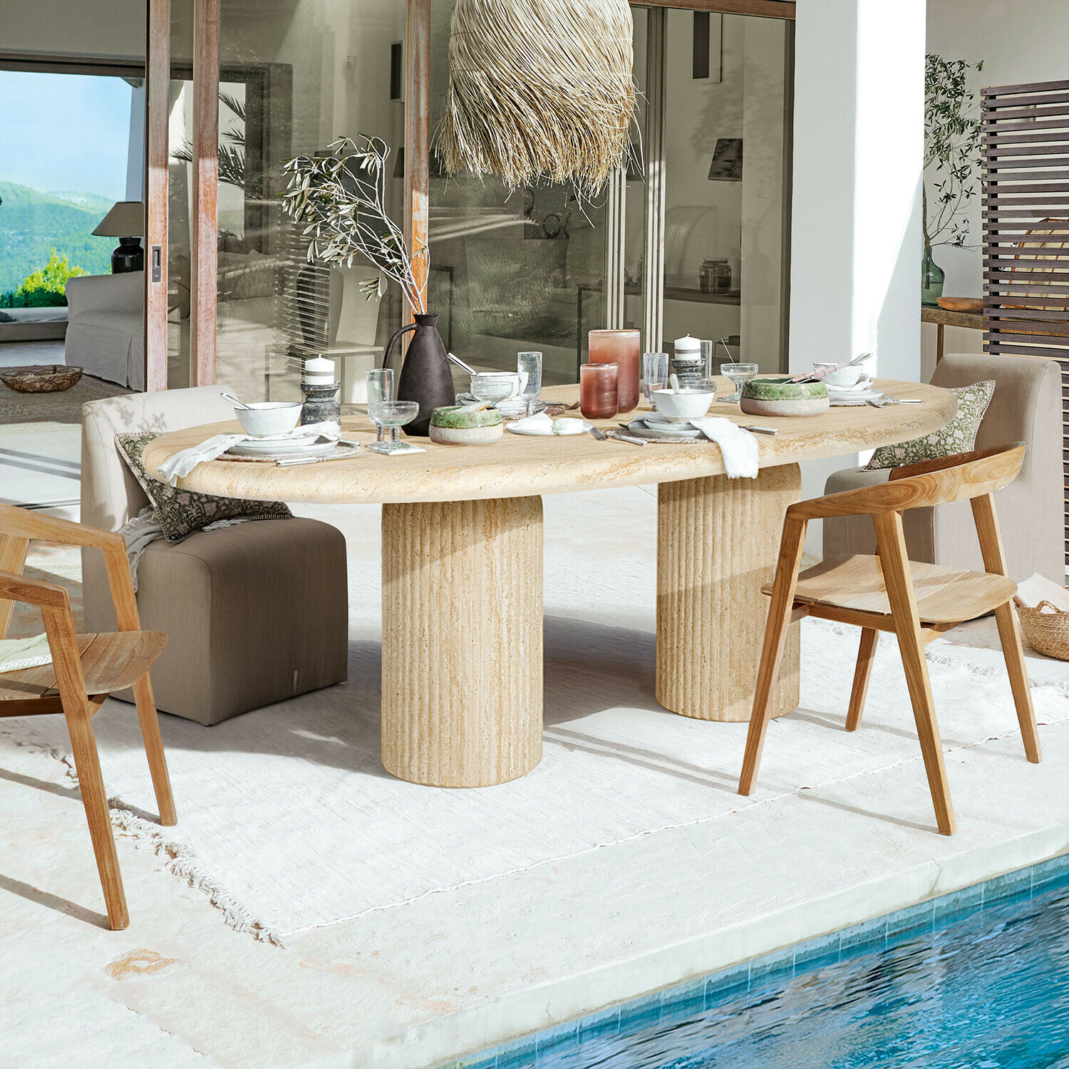 Mesa Calerio Mesa ovalada de piedra beige con dos patas cilíndricas en terraza junto a piscina con sillas de madera