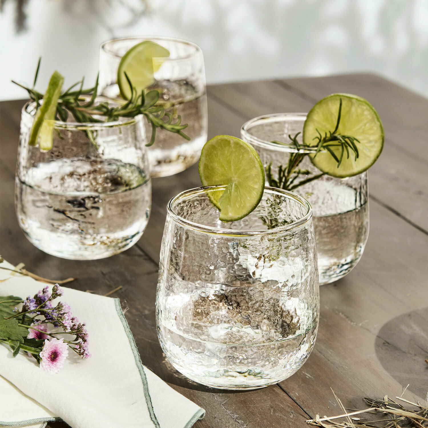 Set de 4 vasos Valters Conjunto de cuatro vasos de vidrio martillado con rodajas de lima y ramitas de romero sobre mesa de madera