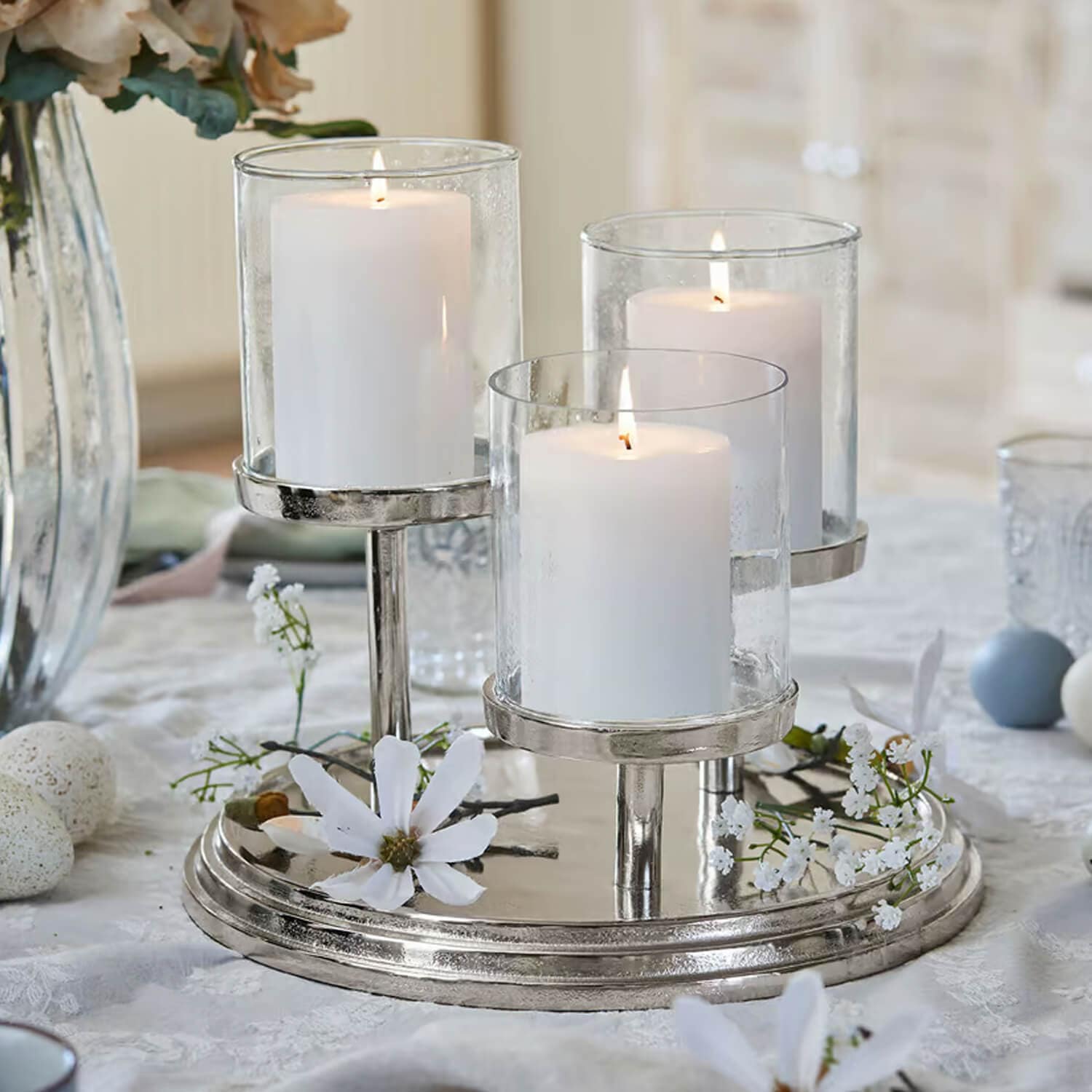Candelabro triple de metal plateado con tres velas blancas y flores decorativas sobre la mesa