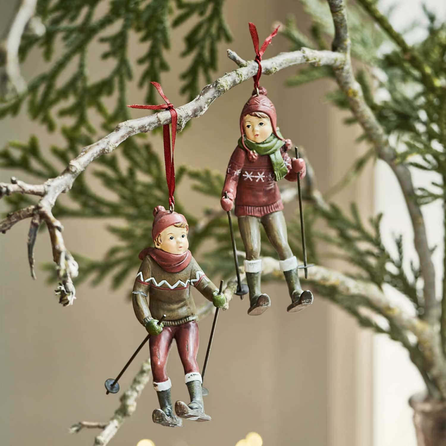Dos adornos colgantes con figuras de niños esquiando con ropa roja y verde en una rama