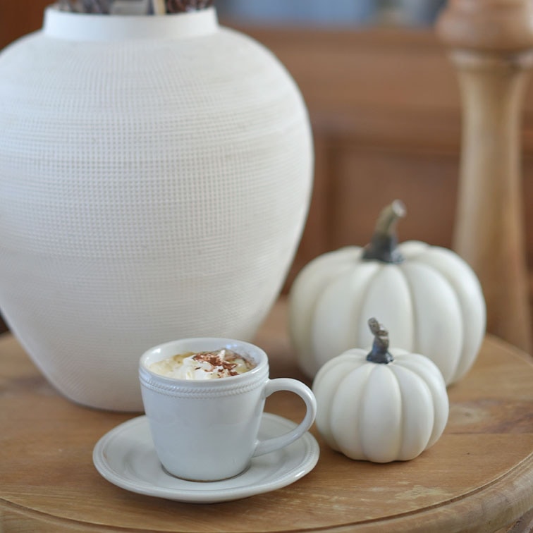 Taza de cerámica blanca con café y nata sobre mesa de madera junto a calabazas decorativas blancas y jarrón