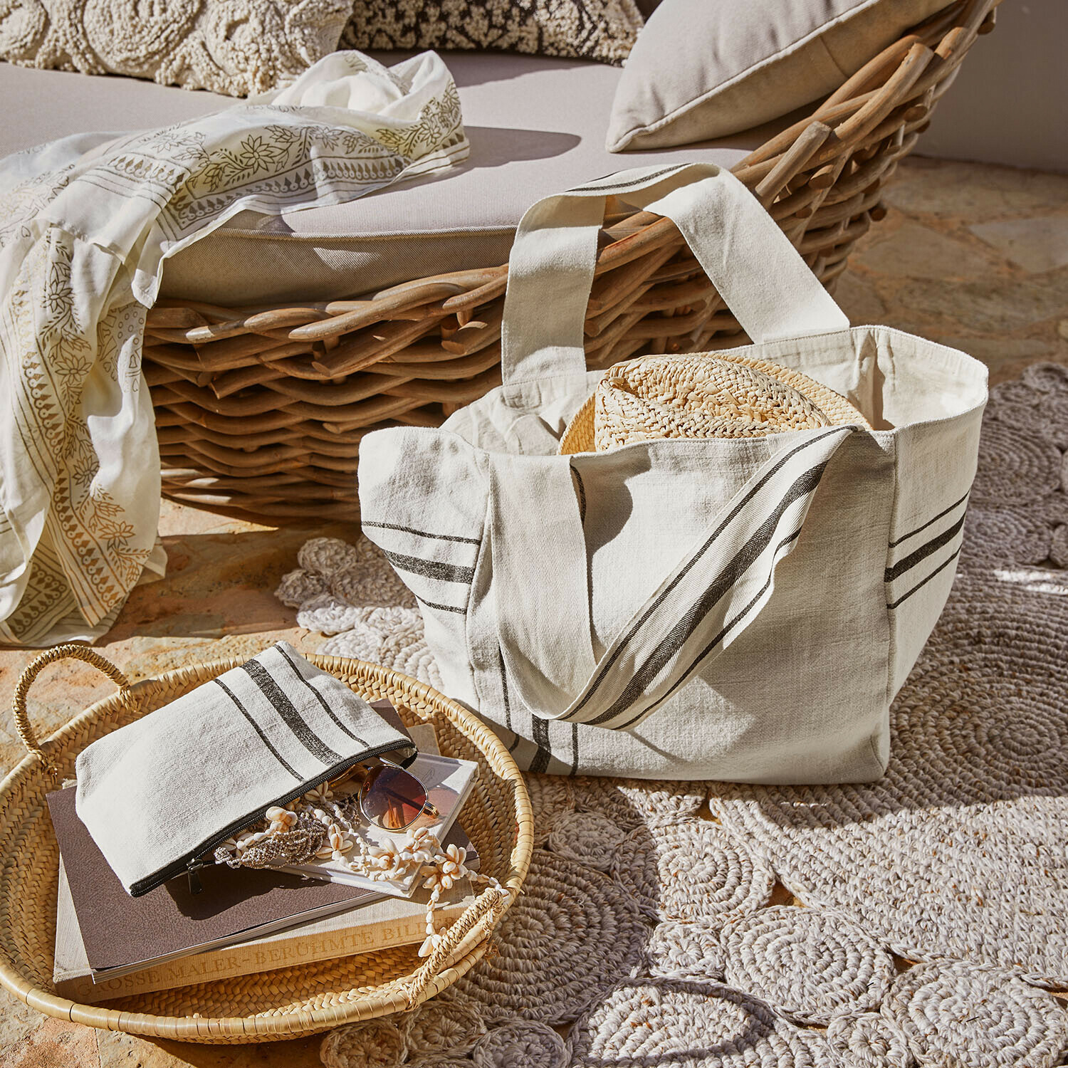 Set de 2 bolsos Bravenda Bolsa de algodón beige con rayas y sombrero de paja sobre sillón de mimbre y alfombra