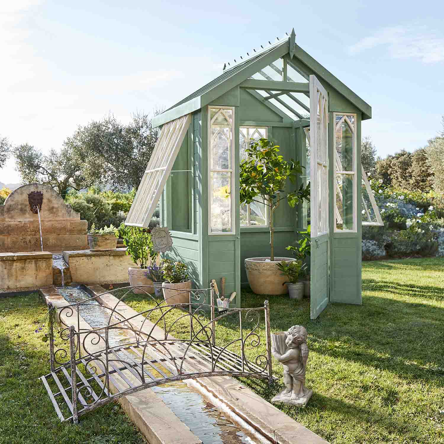 Invernadero de madera verde con ventanas blancas en jardín con plantas y fuente