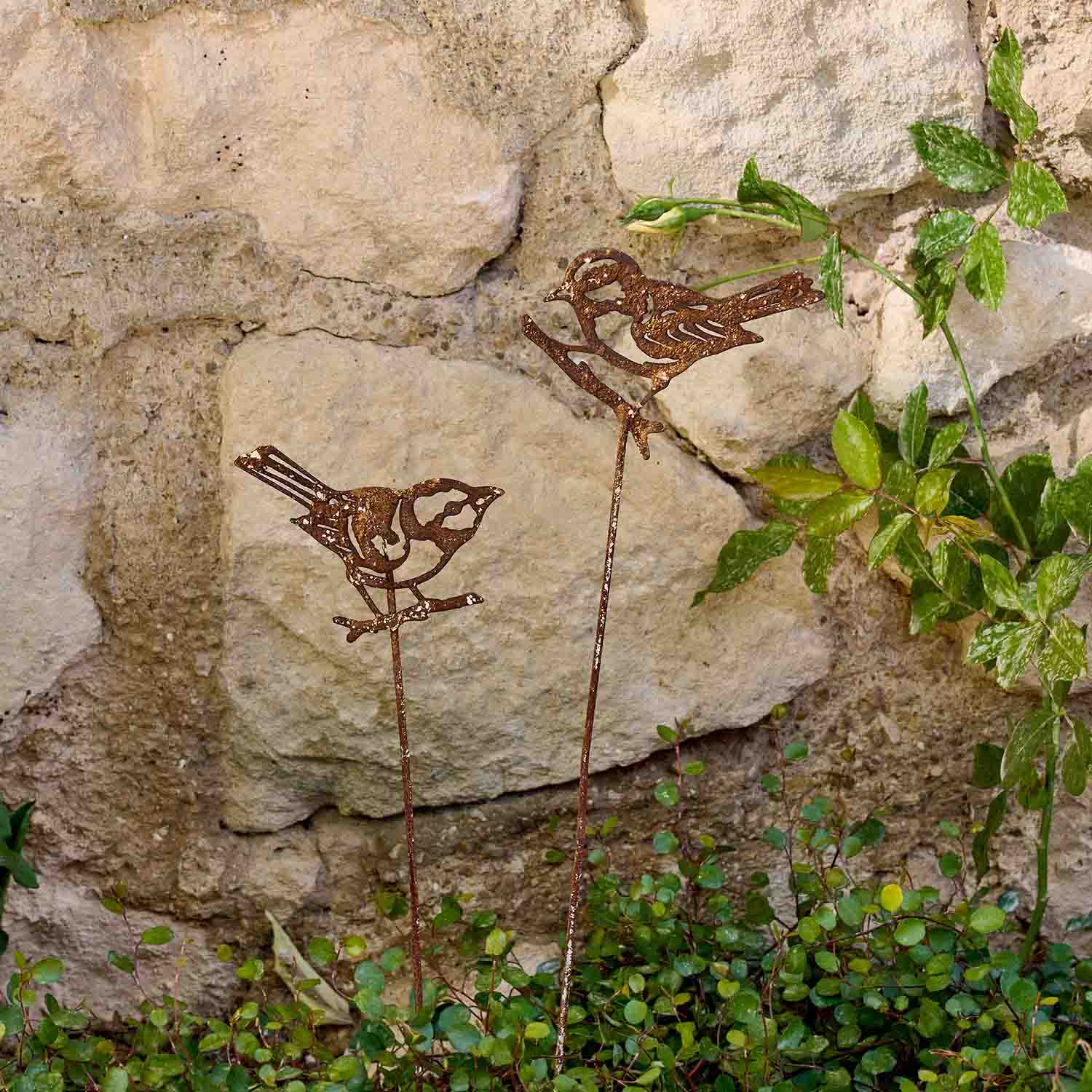 Pájaros decorativos de metal con acabado oxidado en varillas en jardín junto a muro de piedra