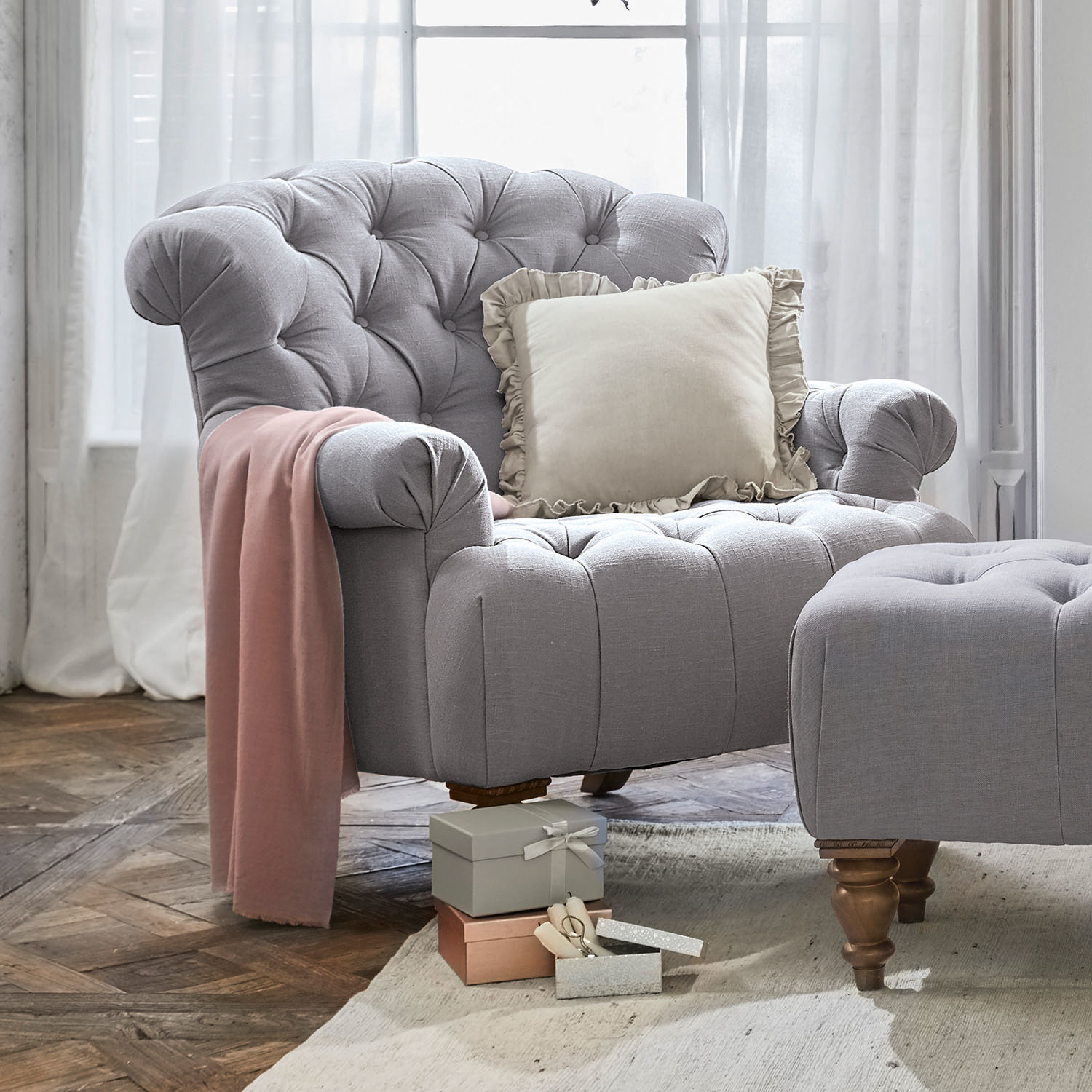 Sillón Springfield Sillón tapizado gris con reposapiés, cojín crema y manta rosa decorativa sobre suelo de madera