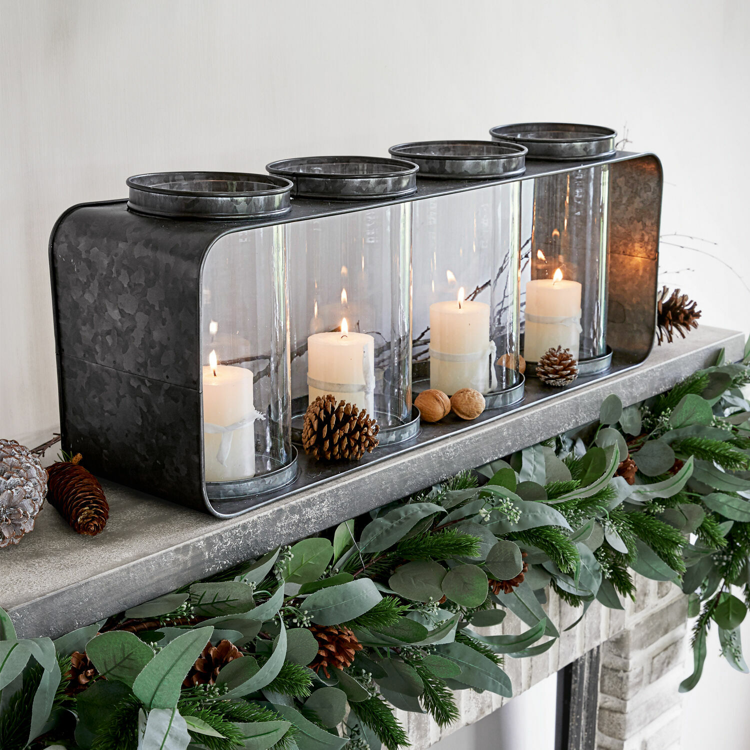 Linterna Sucre Portavelas de metal con cuatro cubiertas de vidrio y velas blancas sobre chimenea decorada con follaje verde