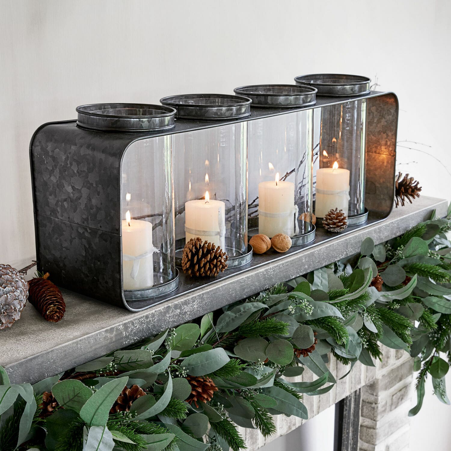 Linterna Sucre Portavelas de metal con cuatro cubiertas de vidrio y velas blancas sobre chimenea decorada con follaje verde