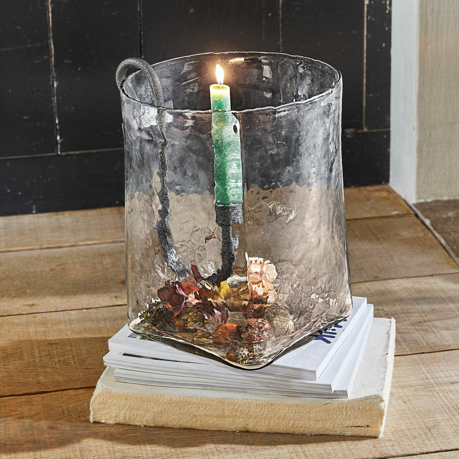 Portavelas de vidrio con asa de metal y vela verde sobre mesa de madera con decoración de piñas
