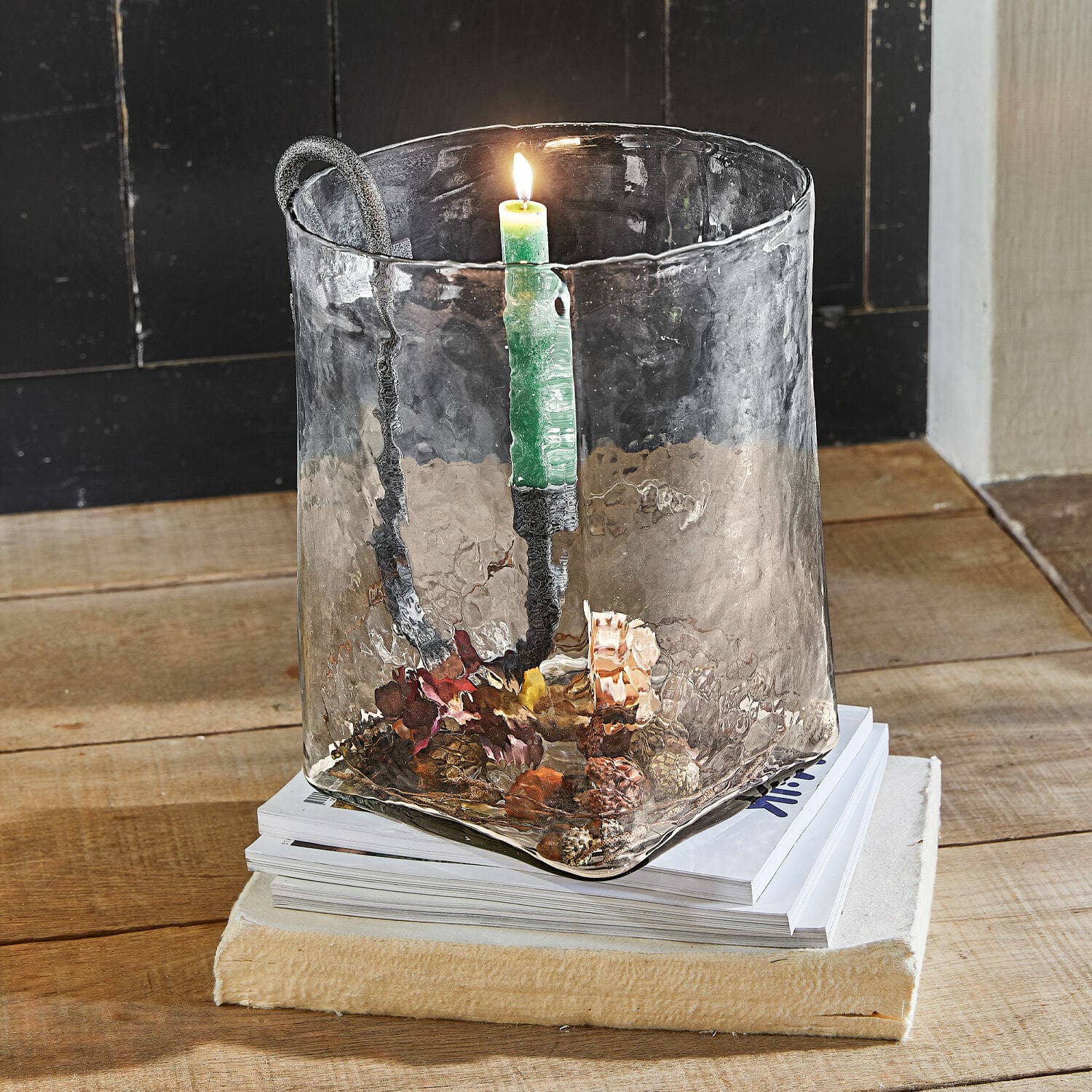 Farolillo Arcene Portavelas de vidrio con asa de metal y vela verde sobre mesa de madera con decoración de piñas