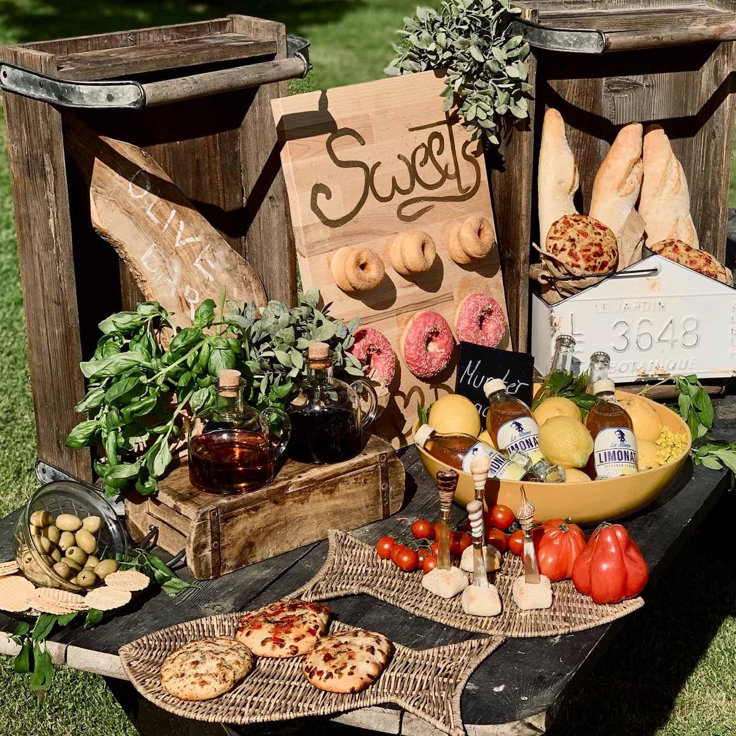 Mesa rústica con cajas de madera, pan, aceitunas, hierbas y limonada al aire libre bajo el sol