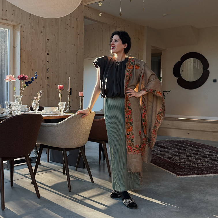 Mujer con falda plisada verde, blusa negra y chal beige con diseño floral en interior moderno