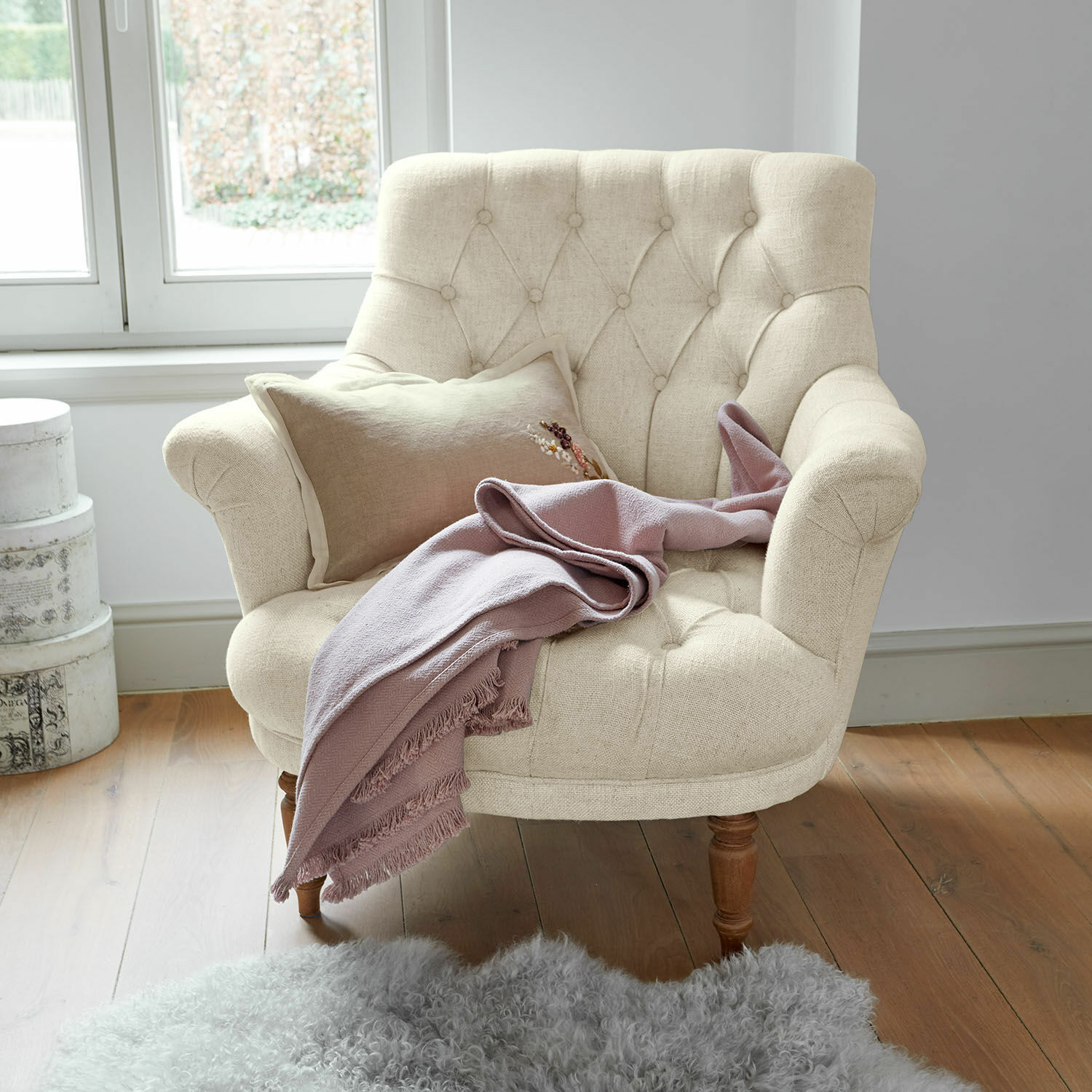 Sillón Rive Dorée Sillón beige tapizado con patas de madera, manta rosa y alfombra de piel de oveja en suelo de madera