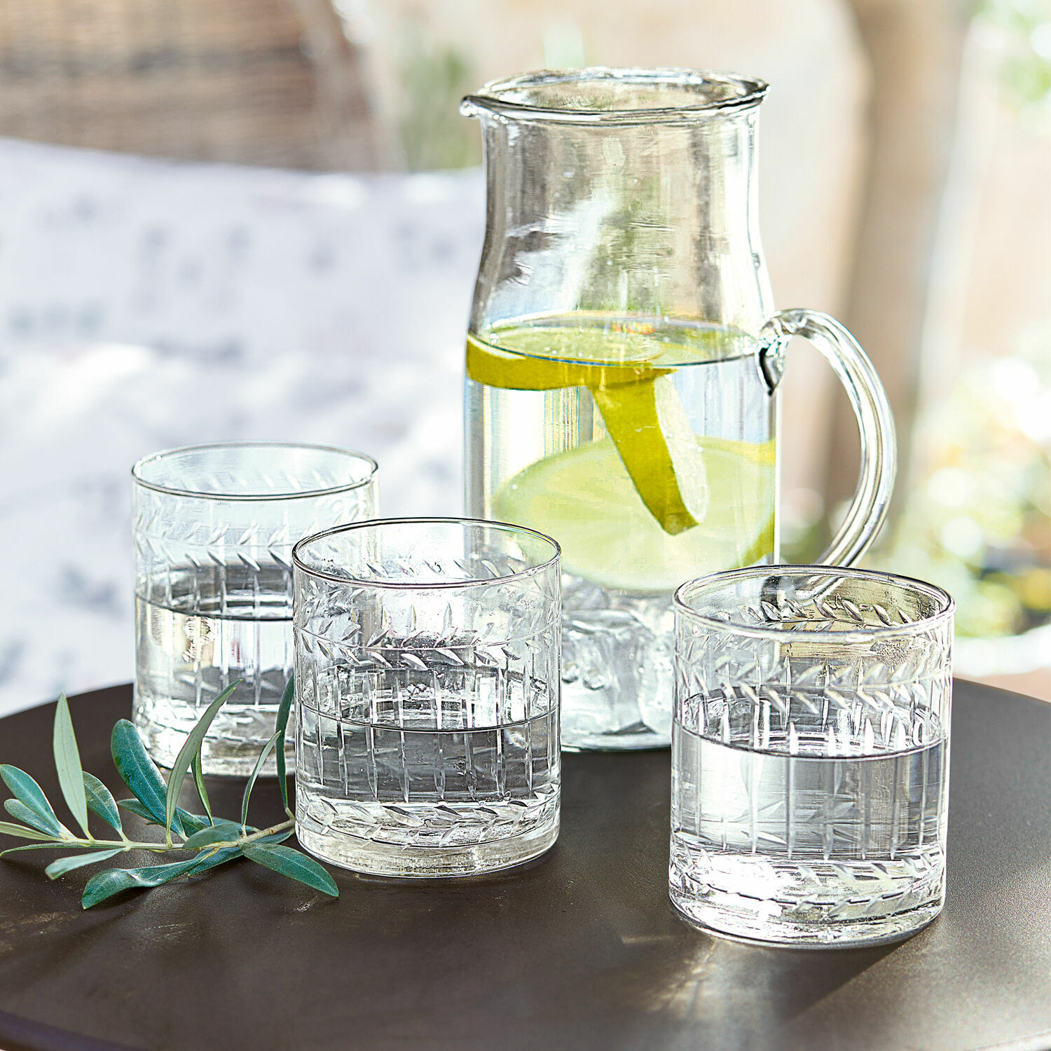 Set de 6 vasos Padiès Jarra de vidrio con rodajas de limón y tres vasos de agua sobre mesa oscura con hojas verdes decorativas