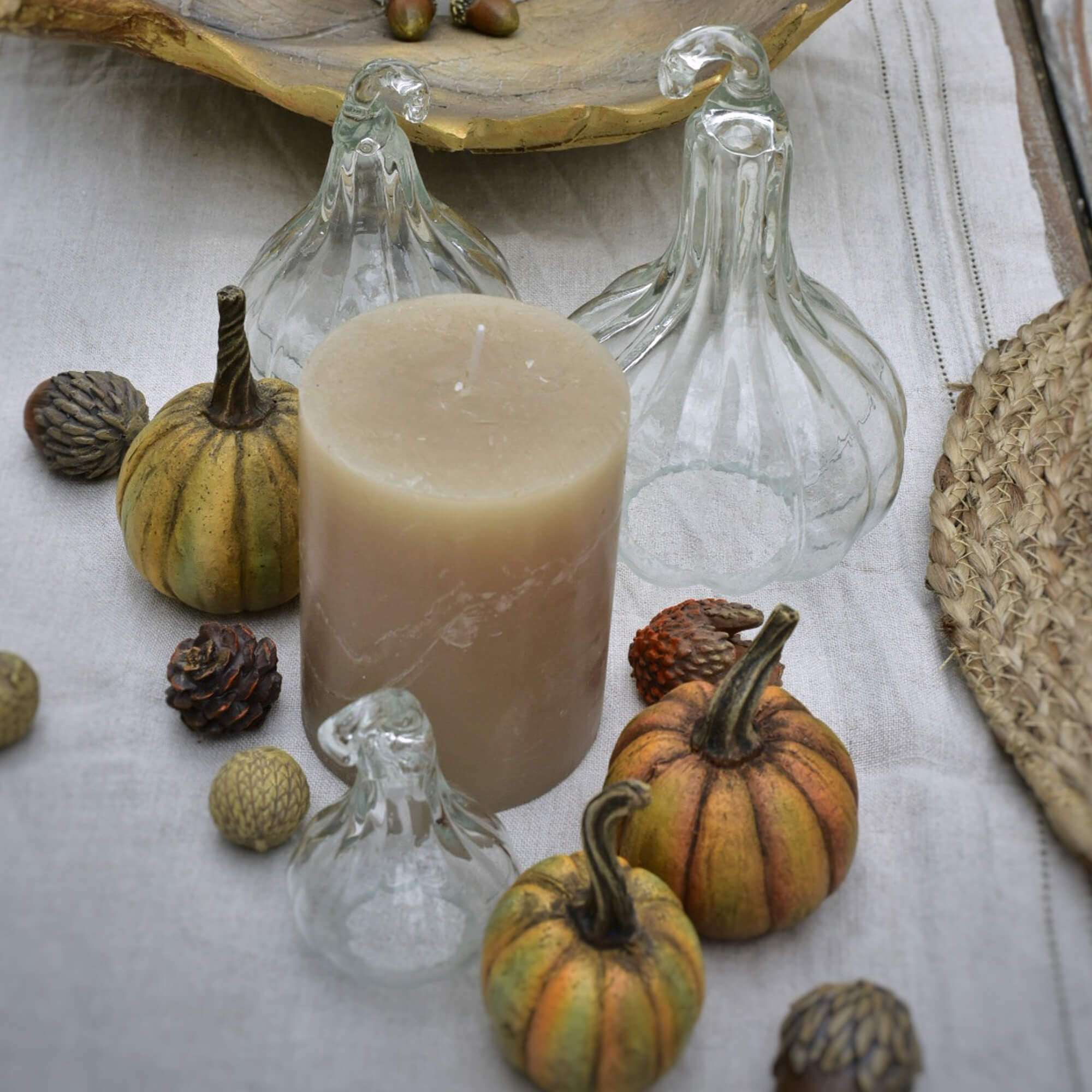 Vela beige con calabazas de cristal y pequeñas decoraciones sobre mantel de lino