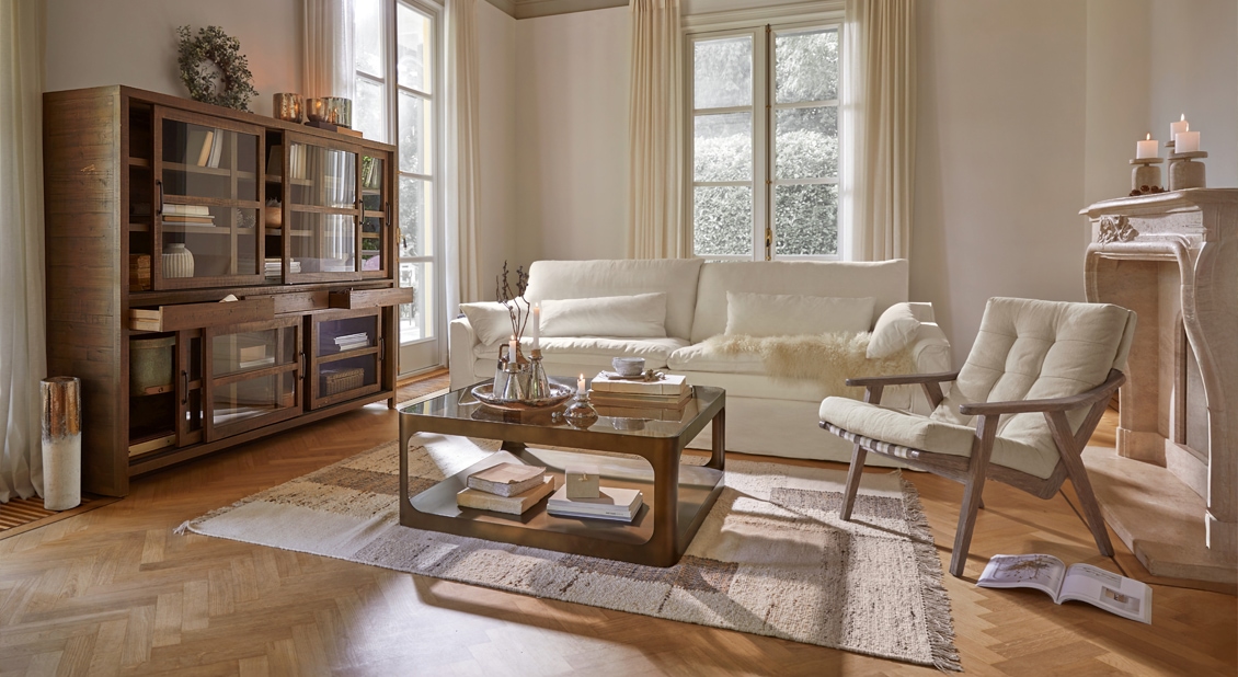 Salón acogedor con sofá crema, sillón de madera y mesa de centro de cristal sobre alfombra, luz natural