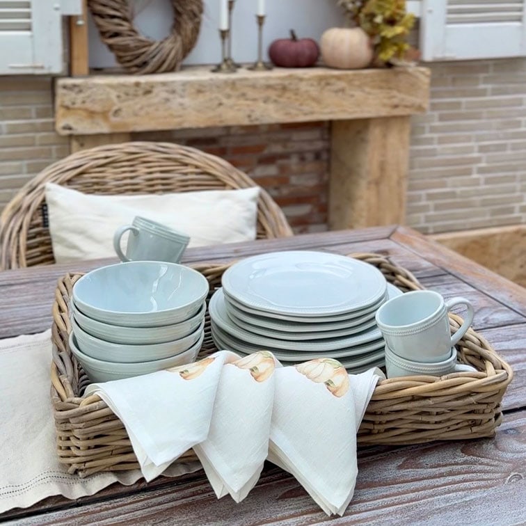 Juego de vajilla de cerámica gris claro con platos, cuencos y tazas en una cesta de mimbre sobre mesa de madera