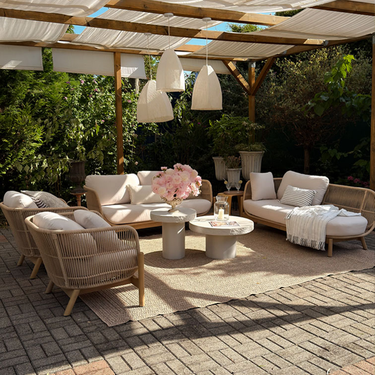 Conjunto de muebles de jardín beige con cojines y estructura de madera bajo pérgola con lámparas colgantes blancas