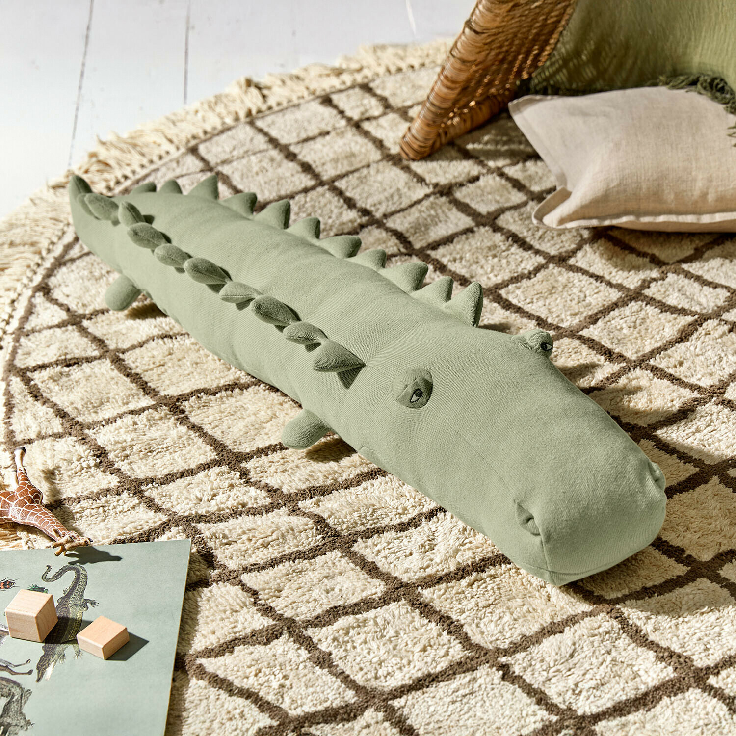 Peluche Zimpy Cojín suave verde con forma de cocodrilo sobre alfombra beige con patrón geométrico