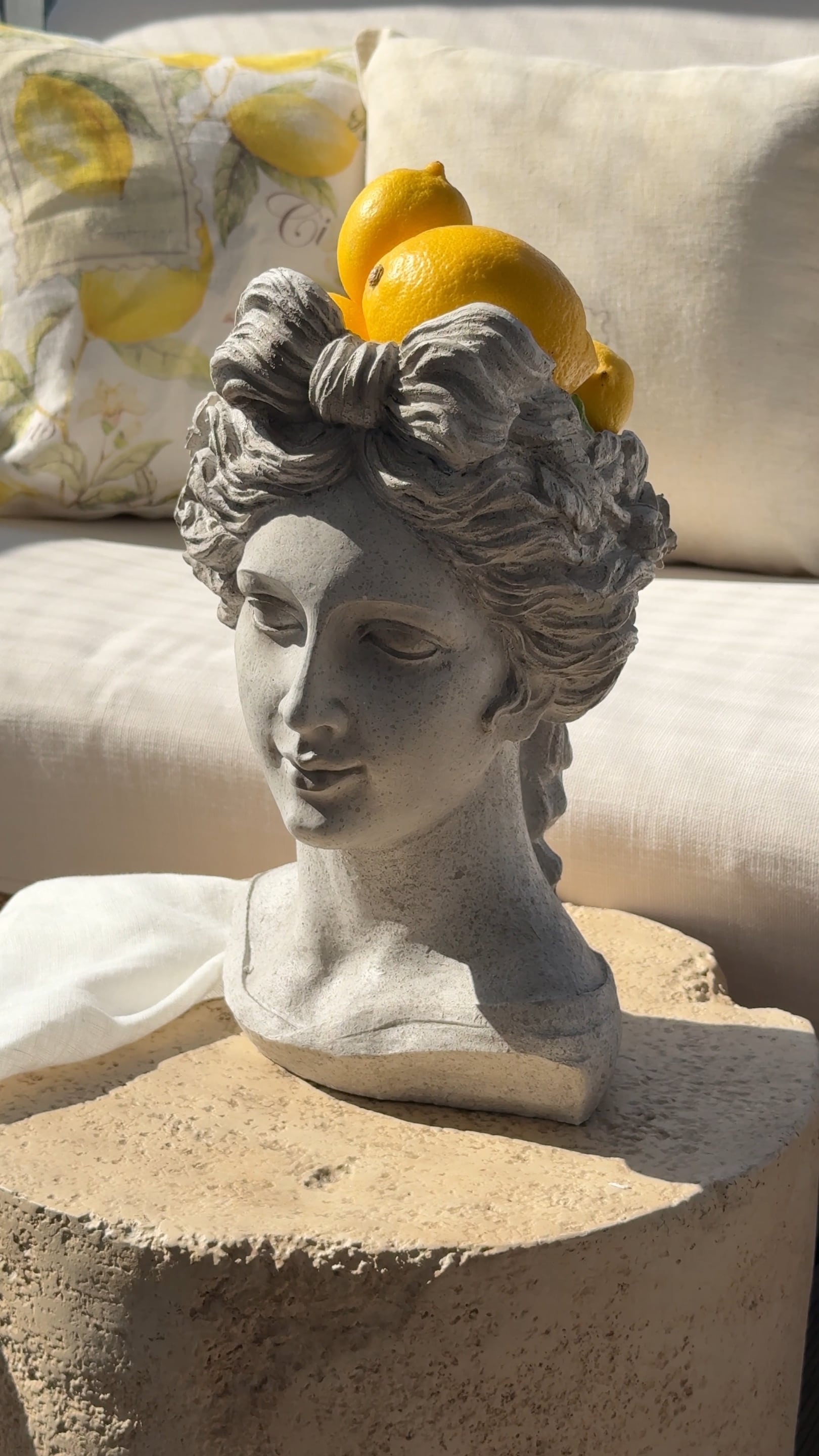 Busto de piedra gris con limones sobre la cabeza frente a cojines claros y tela beige