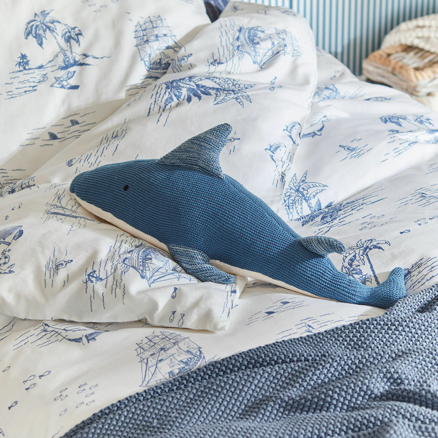 Peluche Cheruboo Cojín azul tejido en forma de tiburón sobre ropa de cama blanca con motivos marinos azules