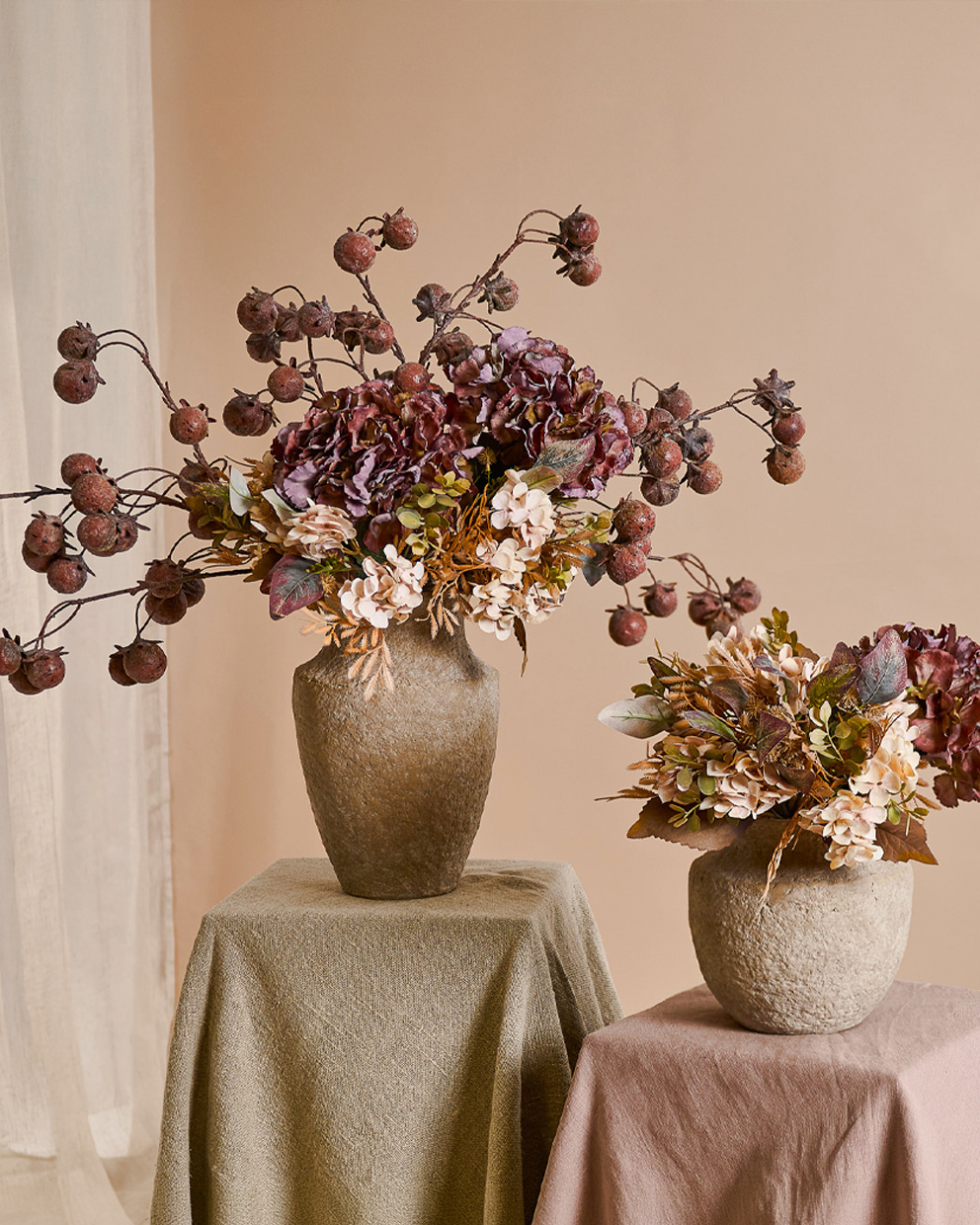 Dos jarrones decorativos de piedra beige con flores otoñales y frutos secos sobre mesas con telas