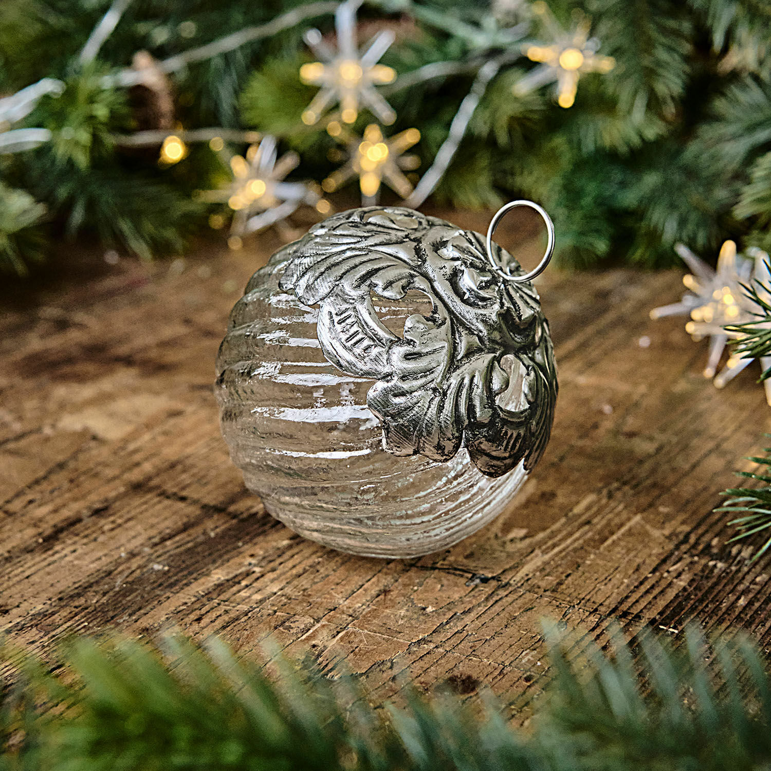 Bola de Navidad Zoriline Bola navideña de vidrio con adorno metálico plateado sobre mesa de madera con ramas verdes