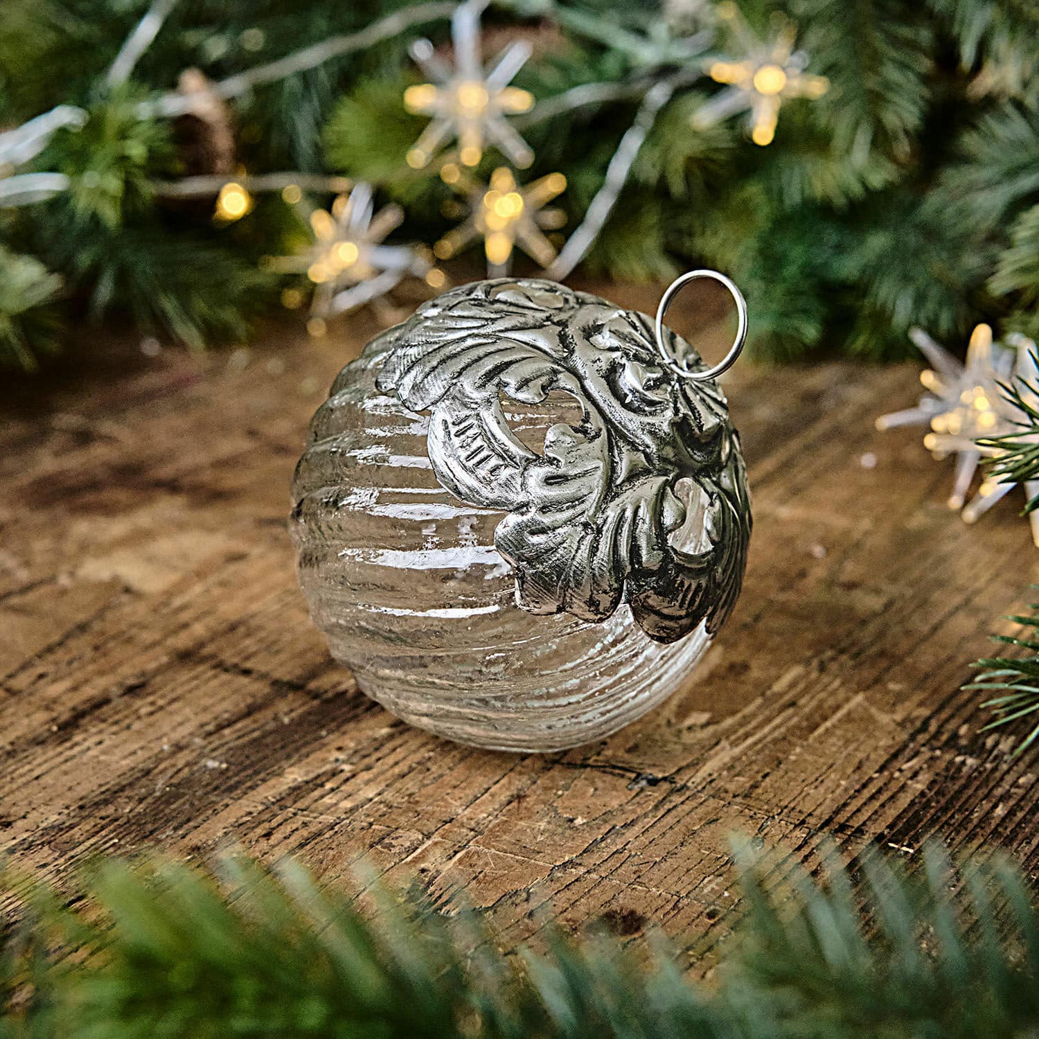 Bola de Navidad Zoriline Bola navideña de vidrio con adorno metálico plateado sobre mesa de madera con ramas verdes