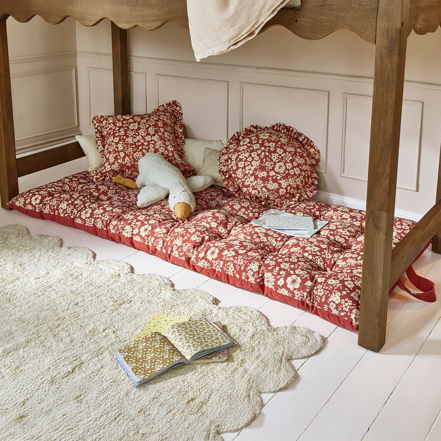 Colchón rojo con estampado floral y cojines bajo una mesa de madera, peluche y libros en el suelo