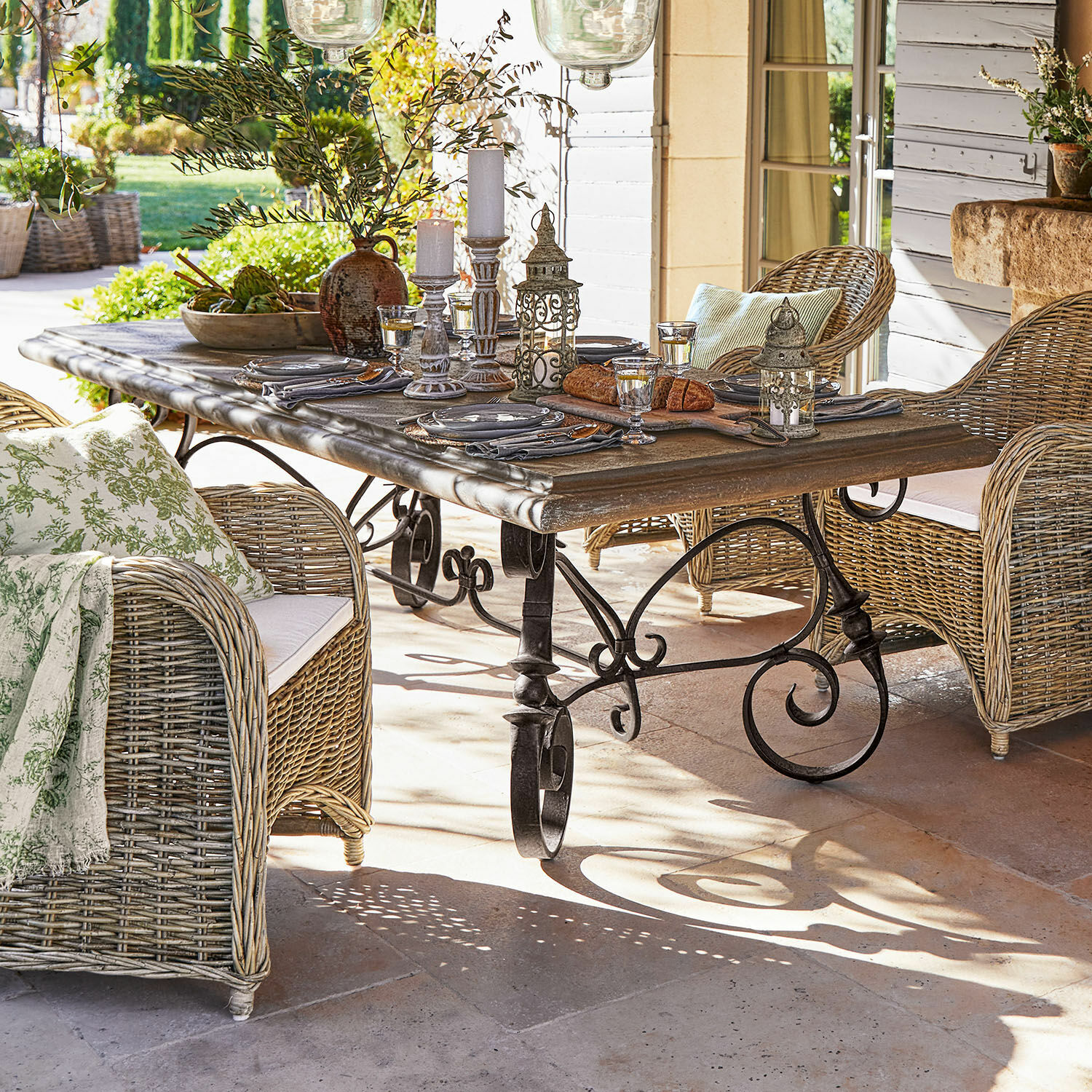 Mesa Bréhéville Mesa de jardín con patas de metal y tablero de madera, rodeada de sillones de mimbre con cojines