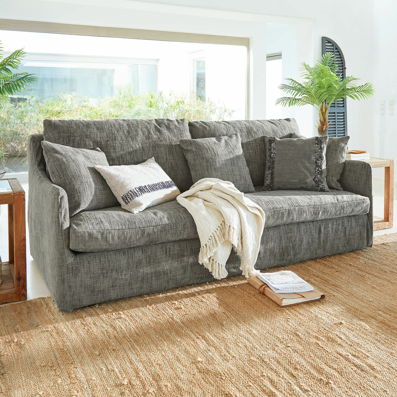 Sofá Shadowfield Sofá gris de tela con cojines y manta crema sobre alfombra natural en sala luminosa