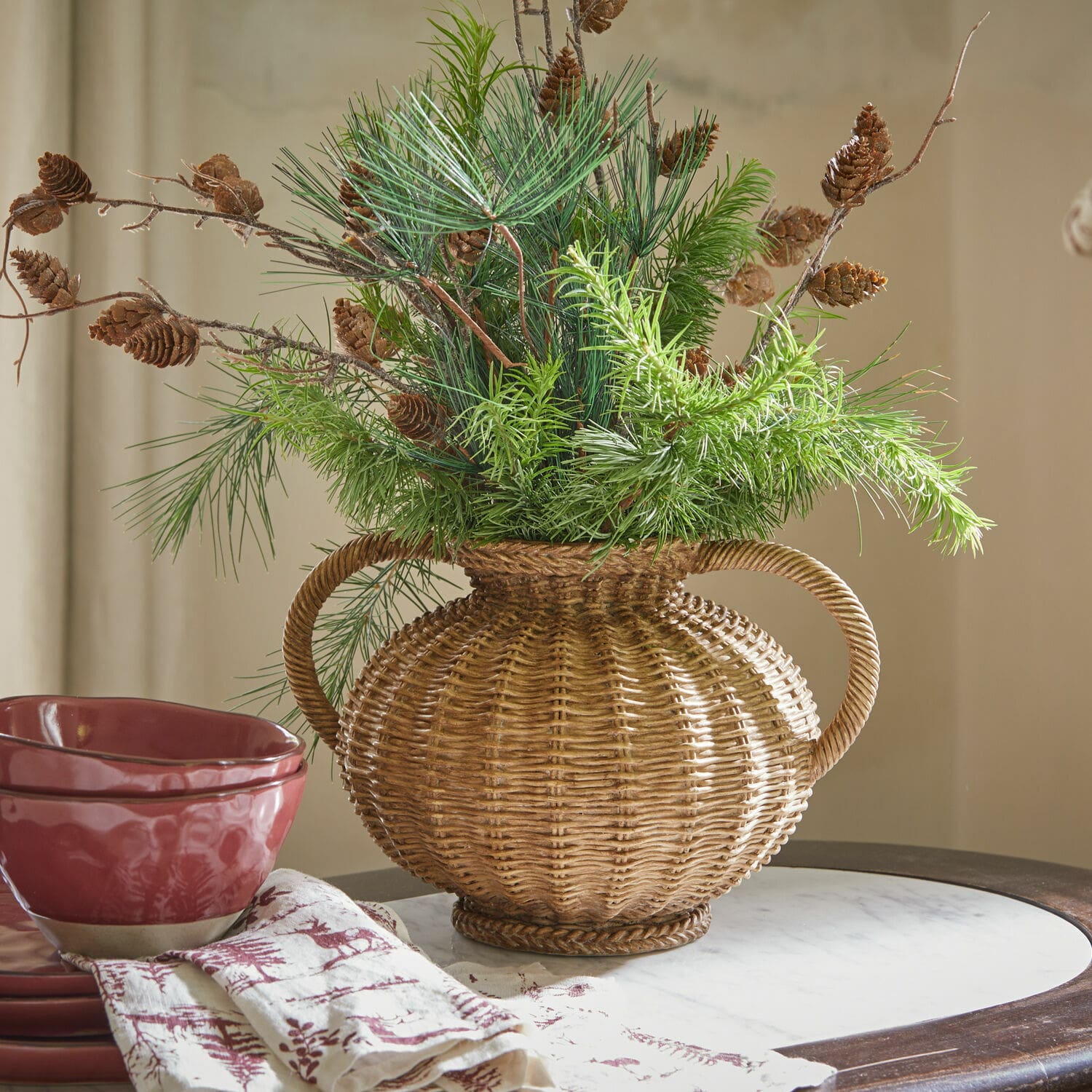 Jarrón Kaelvik Jarrón de mimbre con asas, decorado con ramas verdes y piñas, sobre mesa con vajilla roja