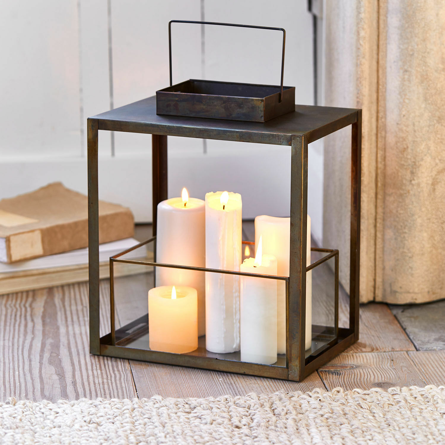Farol Abbracciante Linterna metálica con vidrio transparente y velas blancas encendidas sobre suelo de madera, decoración interior