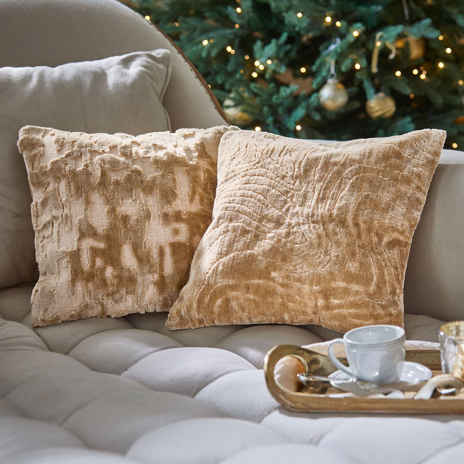 Dos cojines decorativos beige de material suave sobre un sofá claro junto a un árbol de Navidad decorado