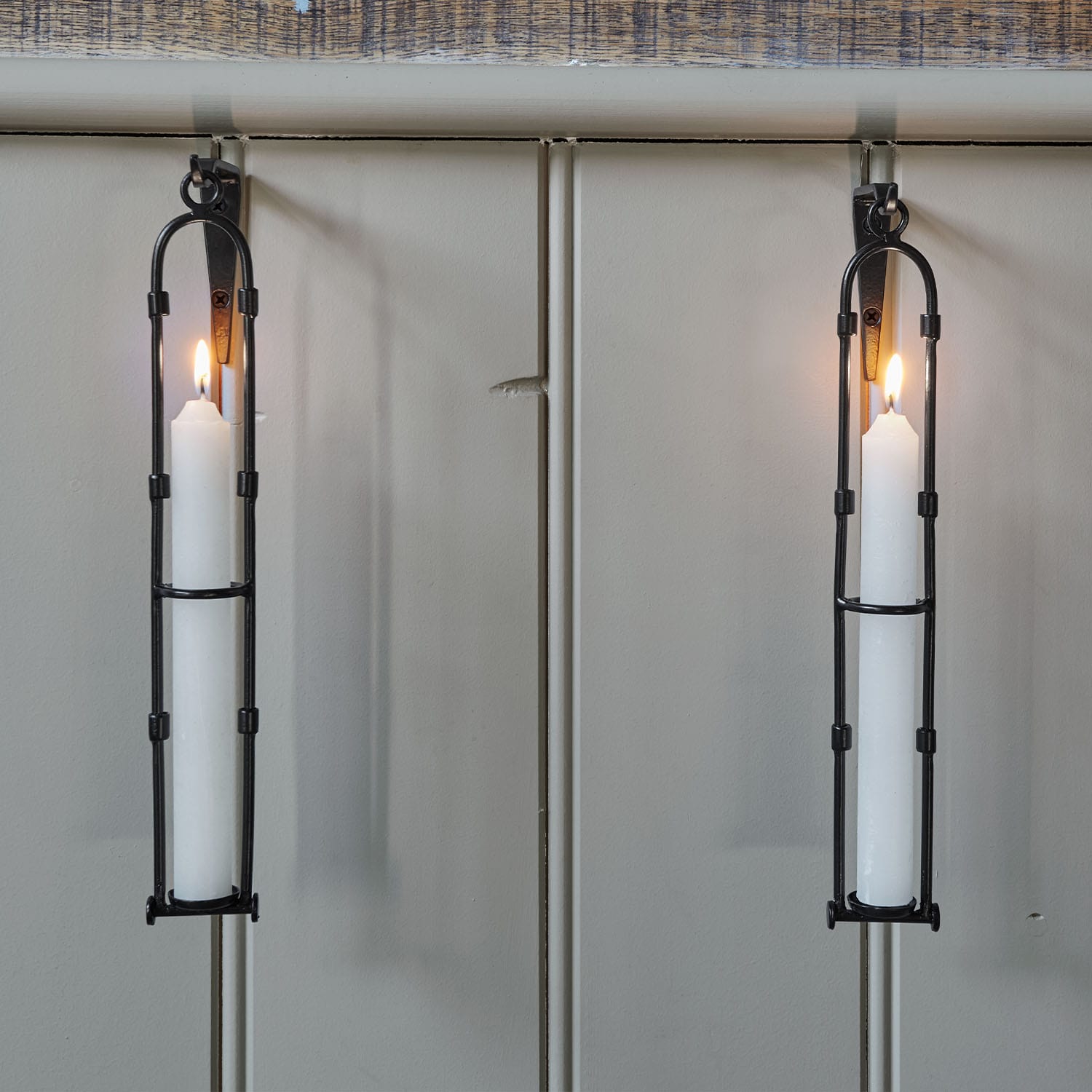Dos candelabros de pared de metal negro con velas blancas encendidas sobre pared gris claro