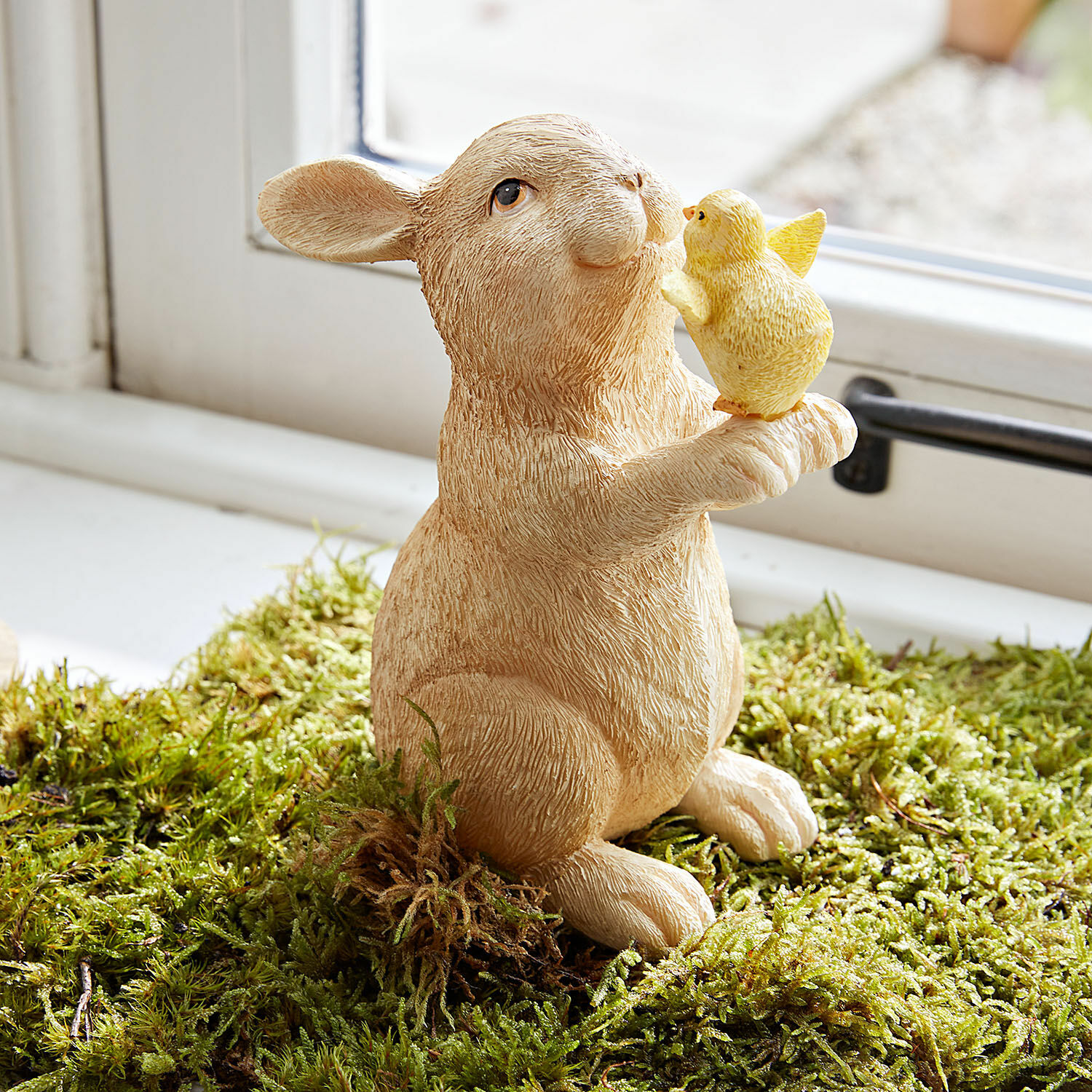 Conejo decorativo Raniah Figura de resina de un conejo beige sosteniendo un pollito amarillo sobre musgo verde, decoración primaveral