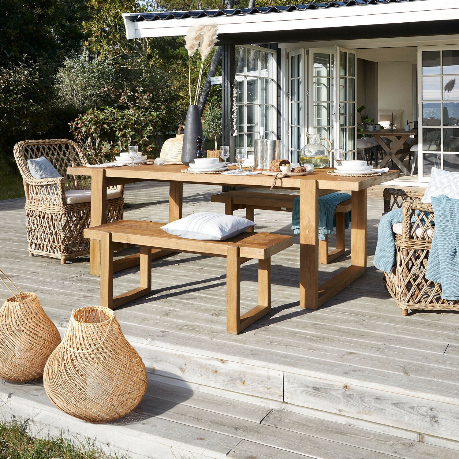 Mesa de jardín de madera con bancos y sillones de mimbre en terraza de madera con decoración natural