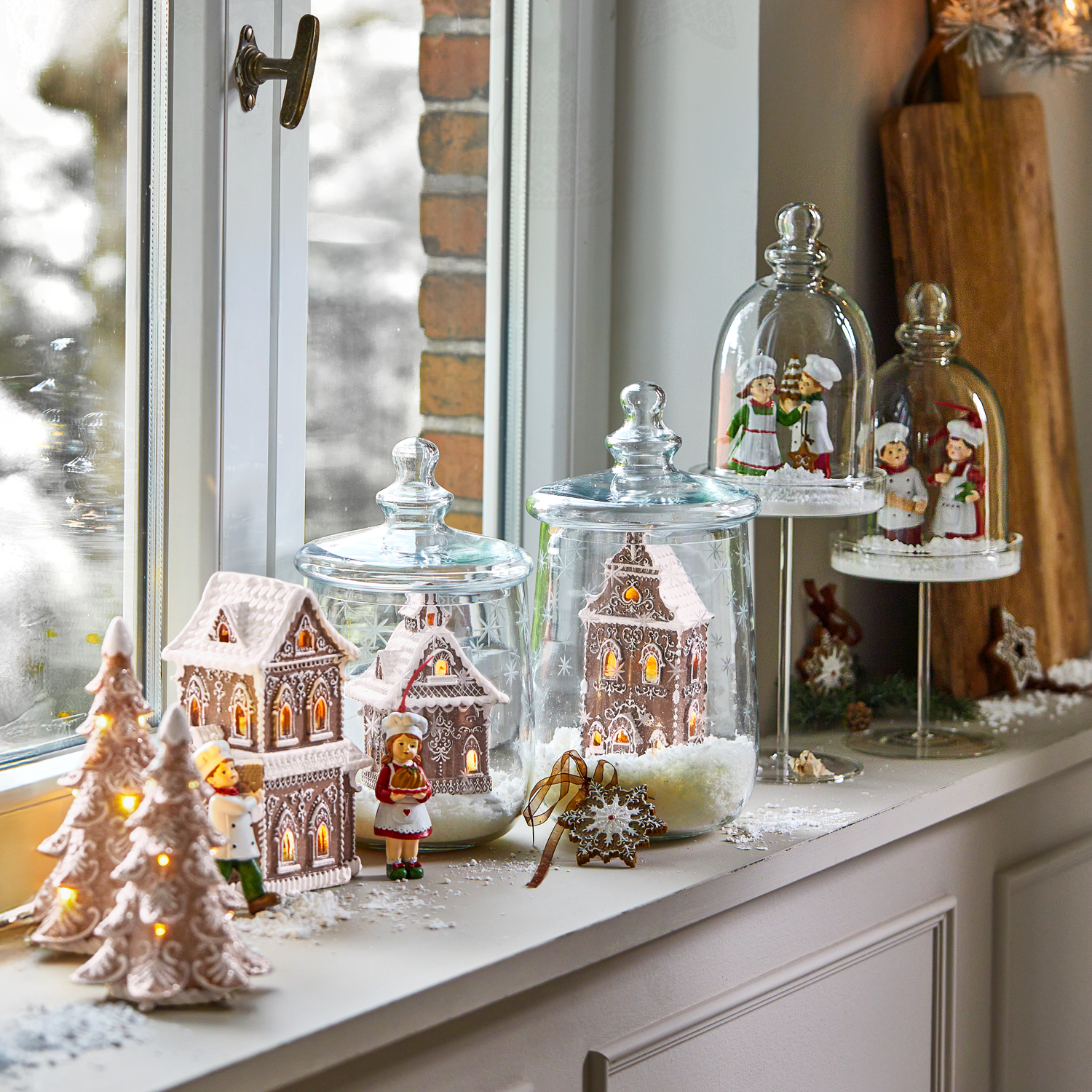 Decoración navideña con casitas de jengibre y figuras bajo campanas de cristal en el alféizar