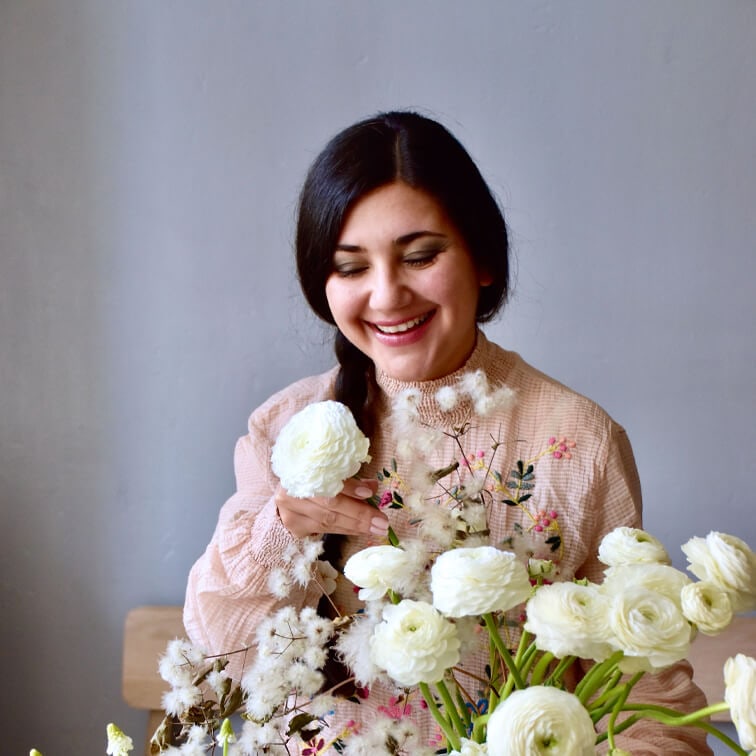 Mujer con blusa rosa bordada sosteniendo flores blancas ranunculus en habitación luminosa