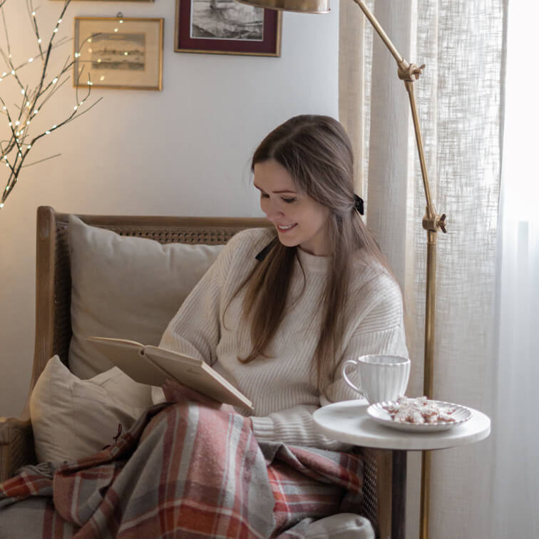 Mujer leyendo un libro en sillón de mimbre con suéter crema y manta cálida a cuadros