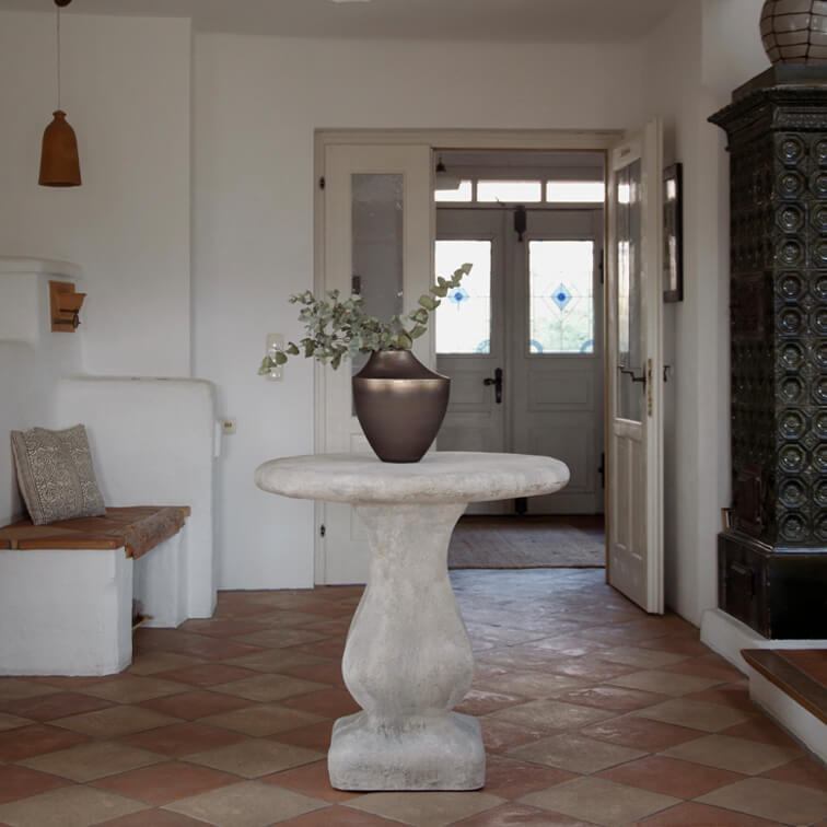 Mesa redonda de hormigón gris claro con jarrón cerámico oscuro y hojas verdes en interior luminoso