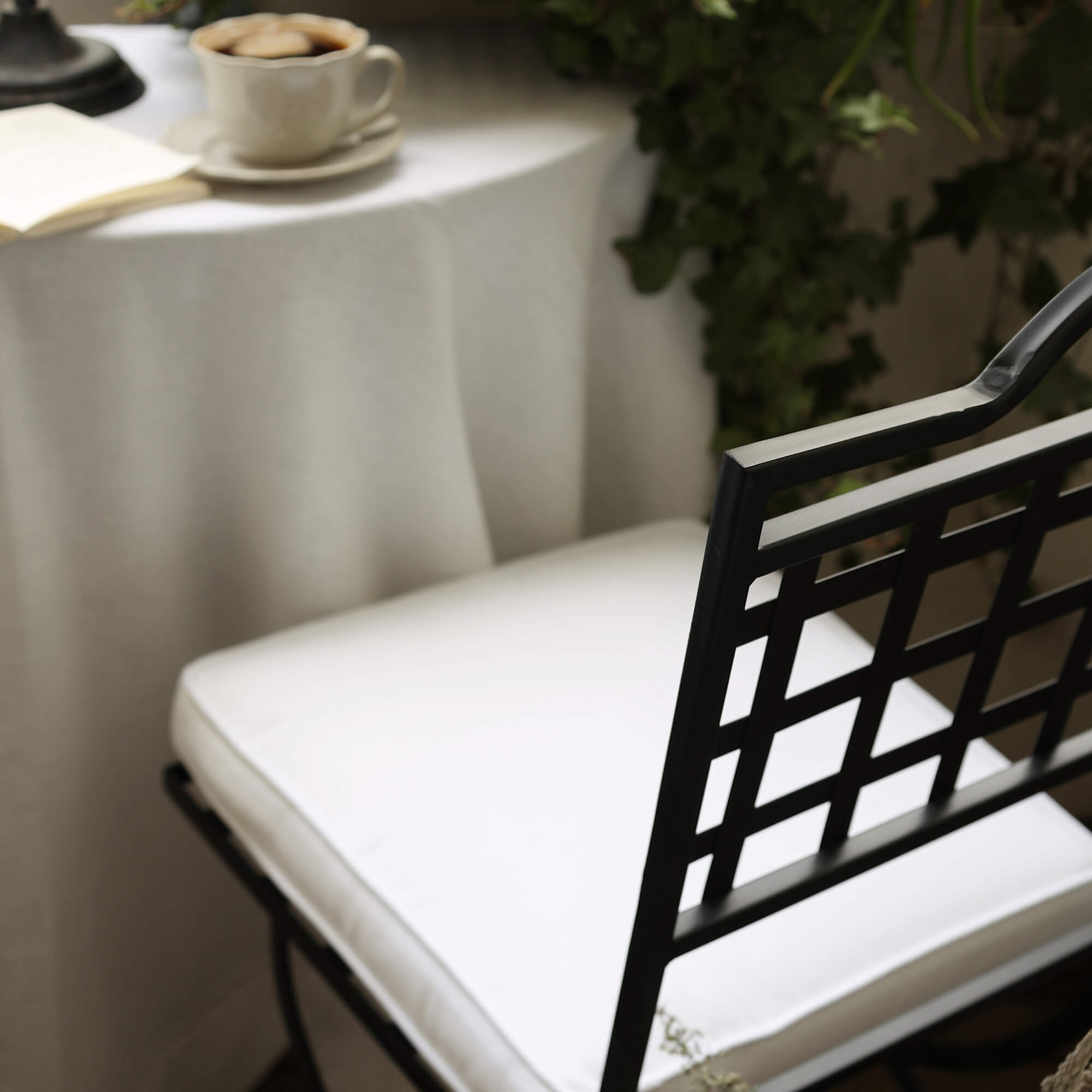 Silla de metal negra con cojín blanco junto a mesa con taza de café y plantas verdes en balcón