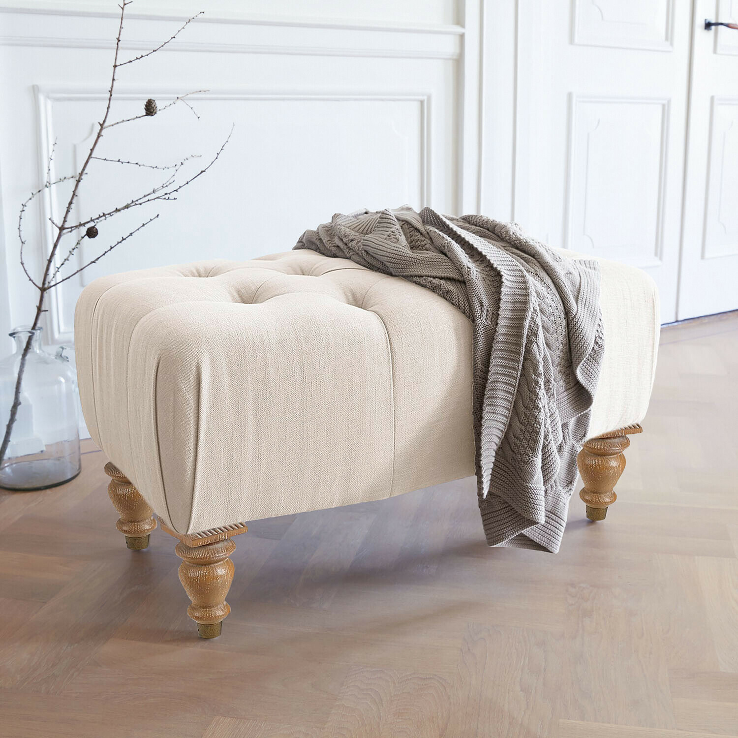 Taburete Durance Puf beige tapizado con patas de madera y manta de punto gris en interior luminoso